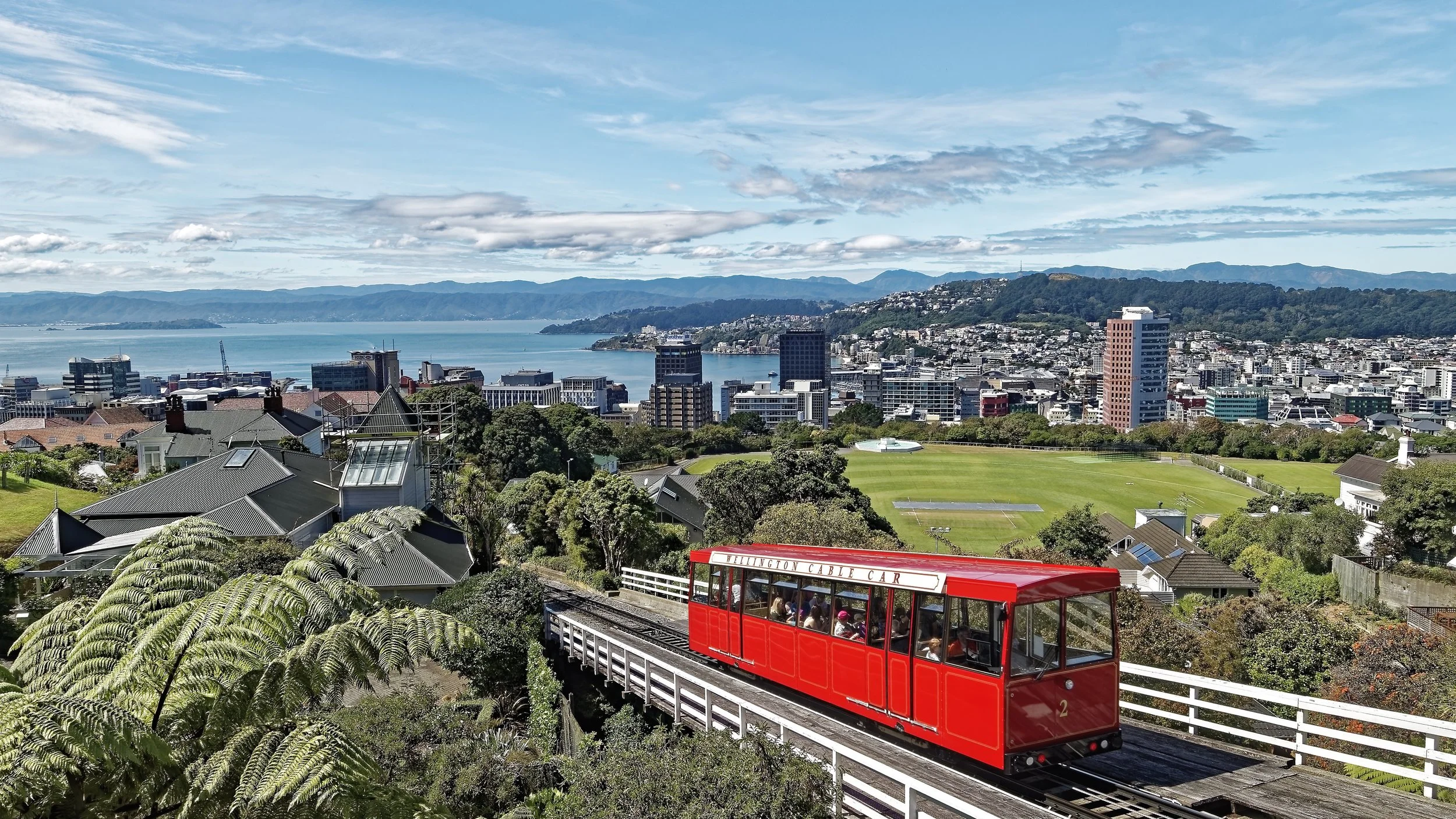 A scenic cityscape overlooking a bay with mountains in the background. In the foreground, a red cable car labeled 'Wellington Cable Car' is traveling along a track through green trees and nearby houses.