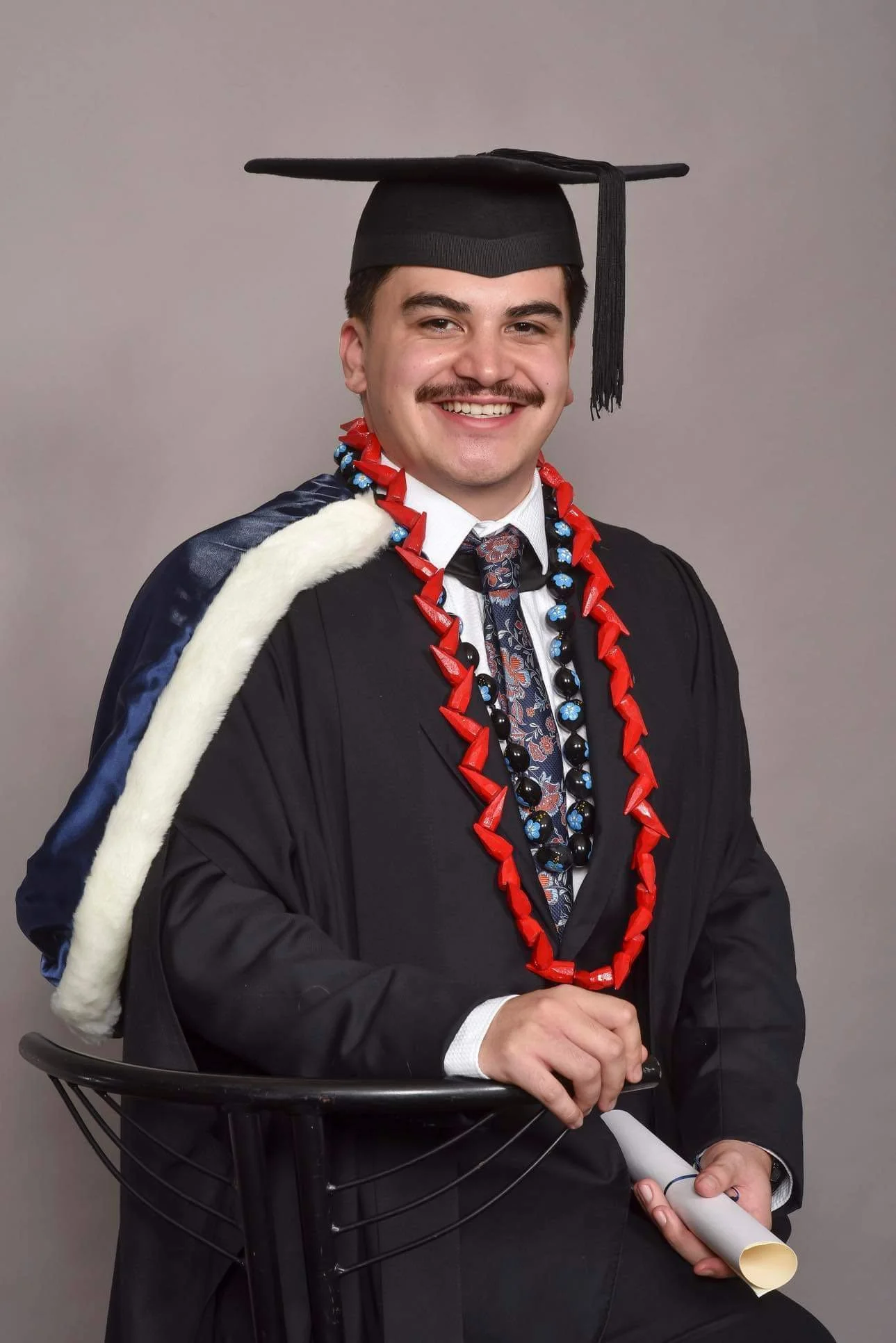 Young man in graduation cap and gown, smiling, with red and blue lei around neck, holding a rolled diploma, seated next to a black chair, with gray background.