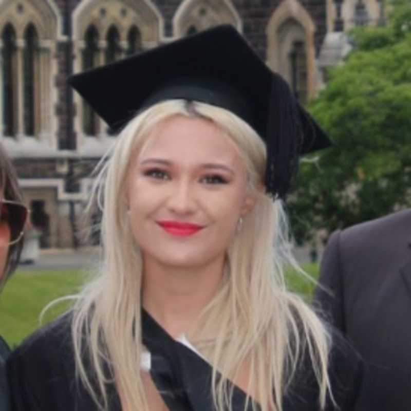 Young woman with blonde hair, wearing a graduation cap and gown, standing outdoors with a historic building and greenery in the background.