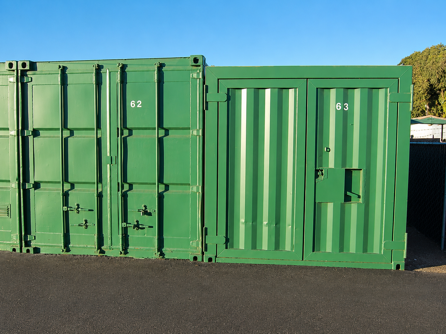 Green shipping containers with numbered doors, set against a clear blue sky and some greenery in the background.