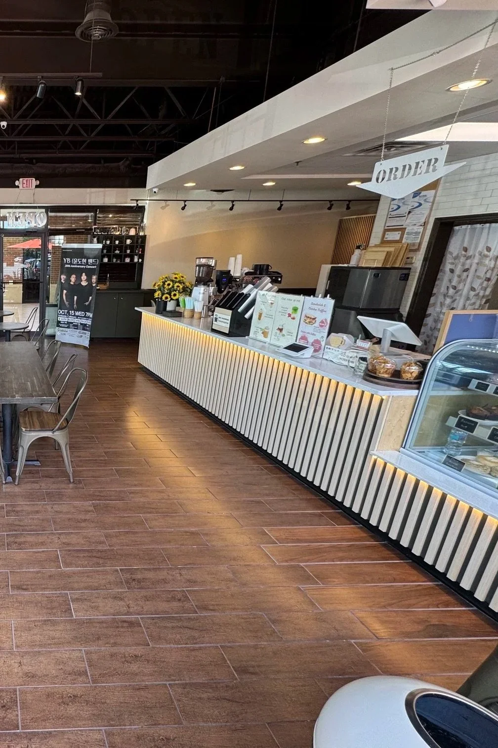 Inside a coffee shop with a modern counter displaying menus, a cash register, and baked goods; seating area with tables and chairs, and a promotional poster on a stand in the background.