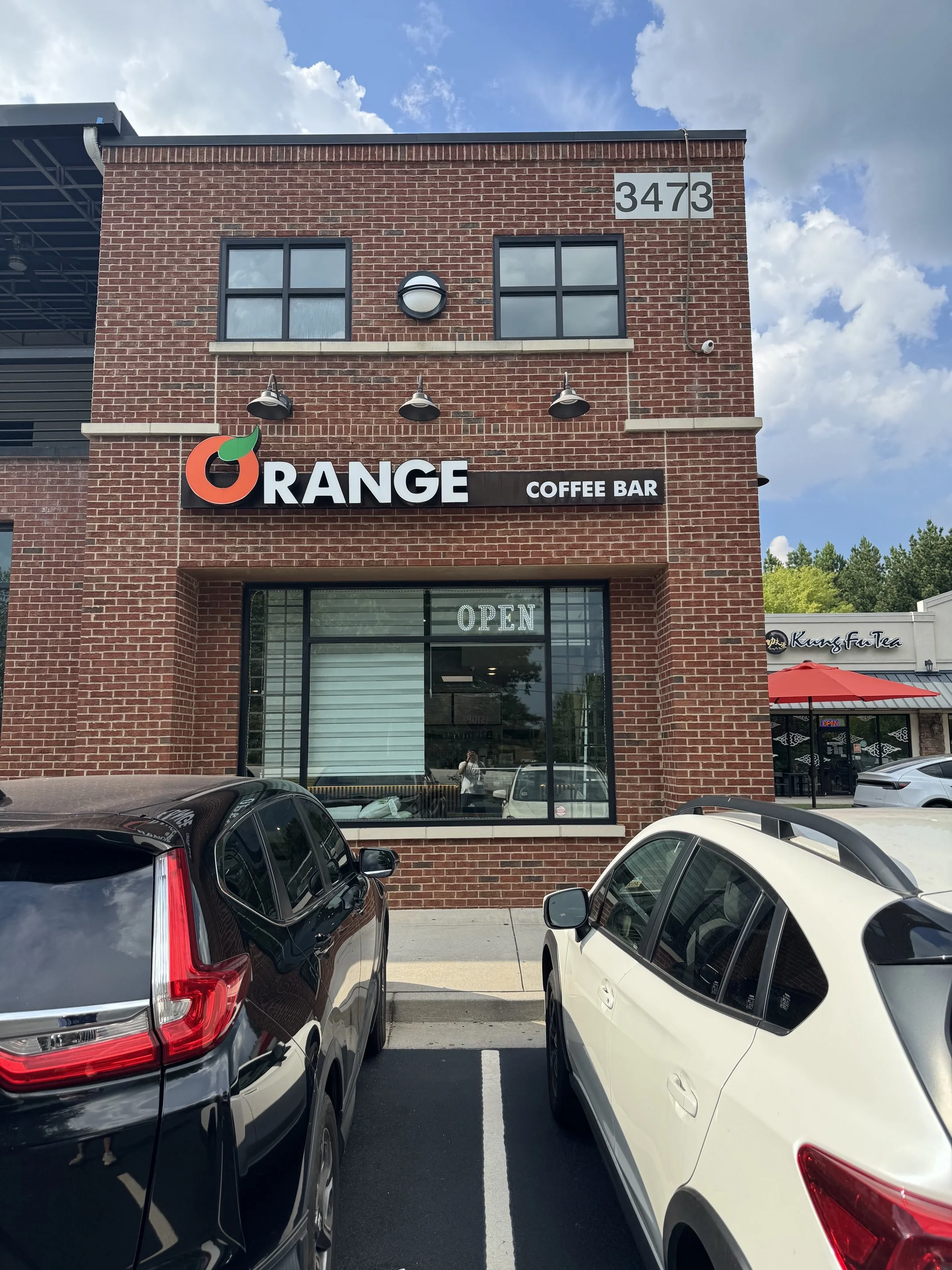 A brick building with a sign that reads 'ORANGE COFFEE BAR' and an 'OPEN' sign in the window. There are cars parked in front of the building, and part of the parking lot and blue sky with clouds are visible.