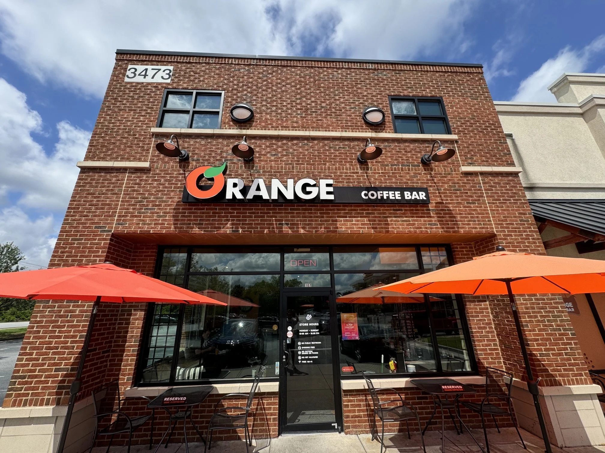 The exterior of a brick coffee shop named 'Orange Coffee Bar' with outdoor seating shaded by an orange umbrella, store hours and policies posted on glass door, and a building number 3473 on the upper wall.