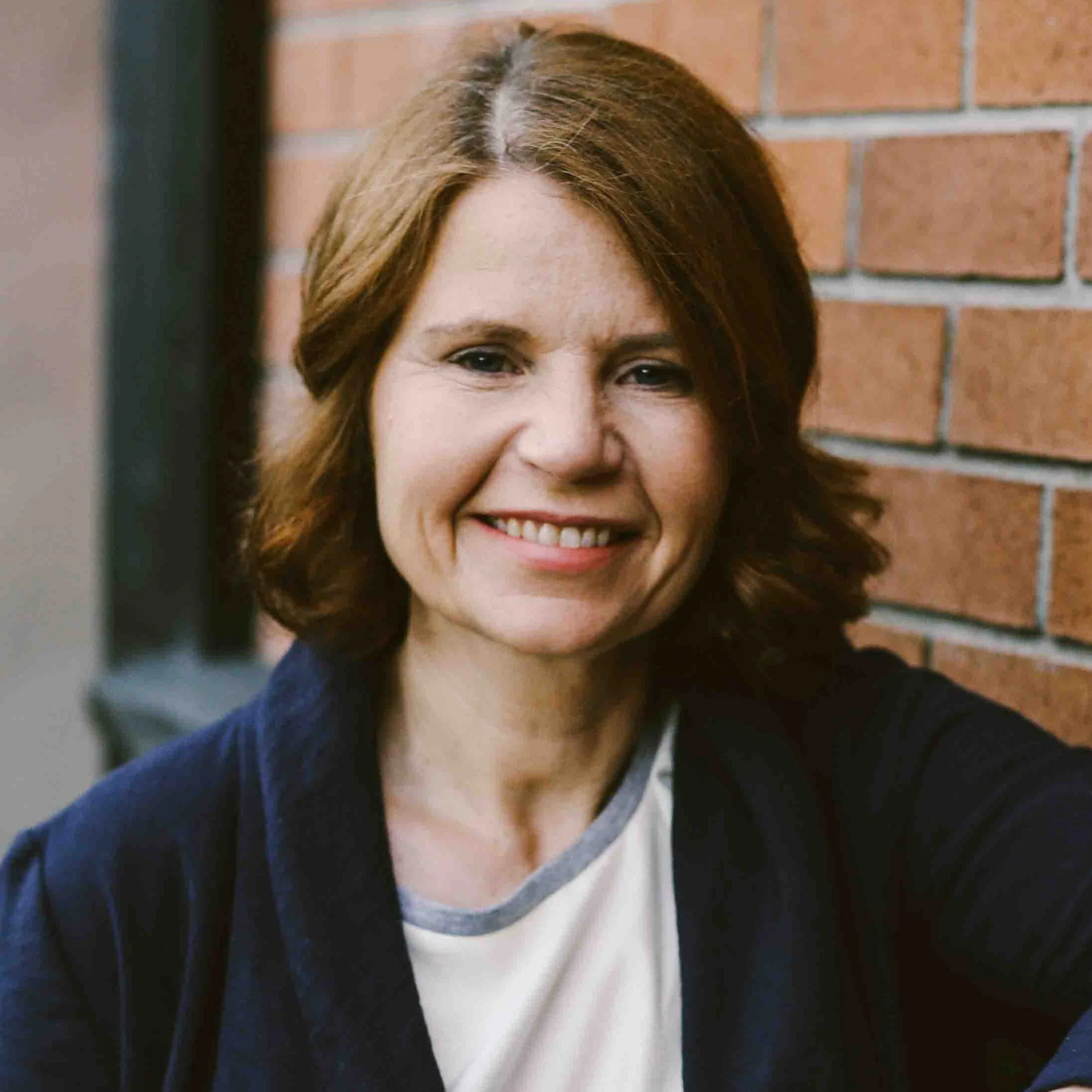 Close-up of a  woman wearing a cream blouse and a navy blazer with light brown hair smiling, a brick wall in the background.