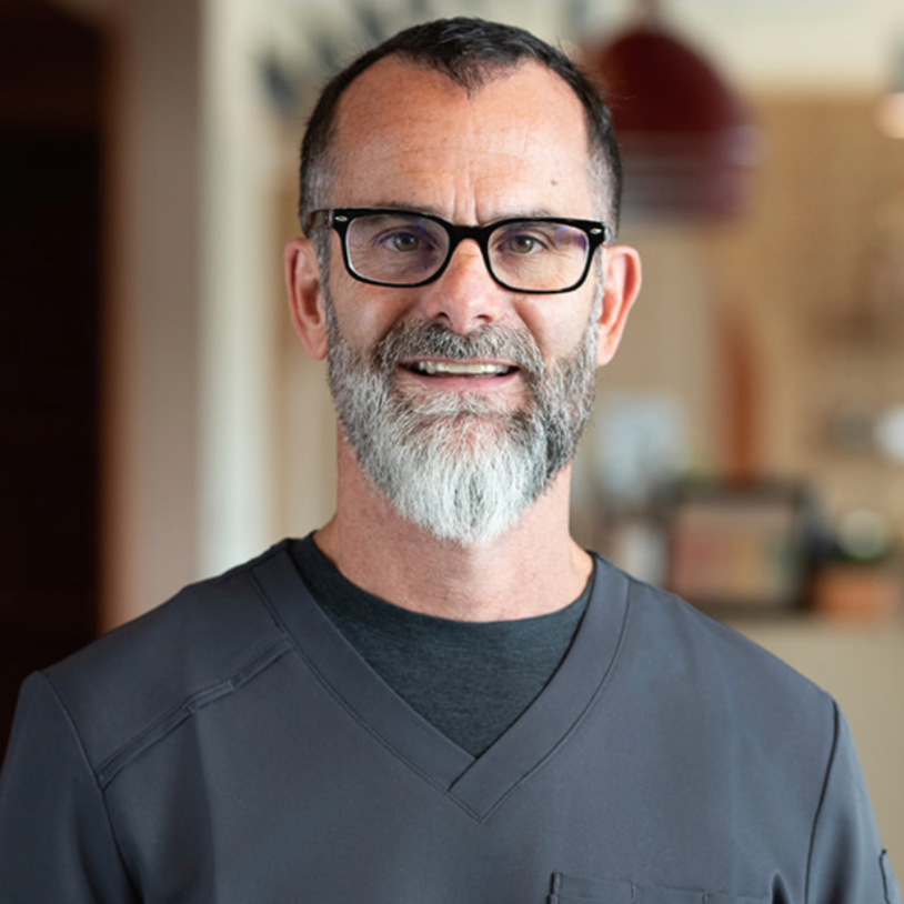 A man with glasses, gray beard, and mustache, smiling at the camera, wearing a dark gray medical scrub top indoors.