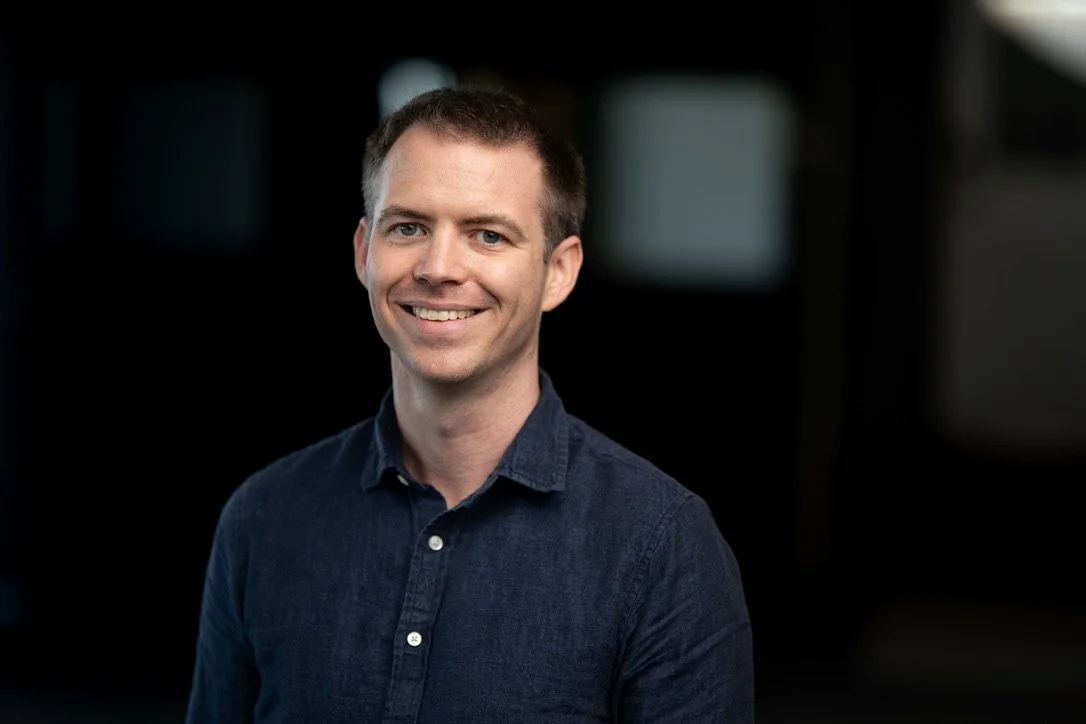 A smiling man with short brown hair, wearing a dark blue button-up shirt, standing against a dark background with blurred windows.