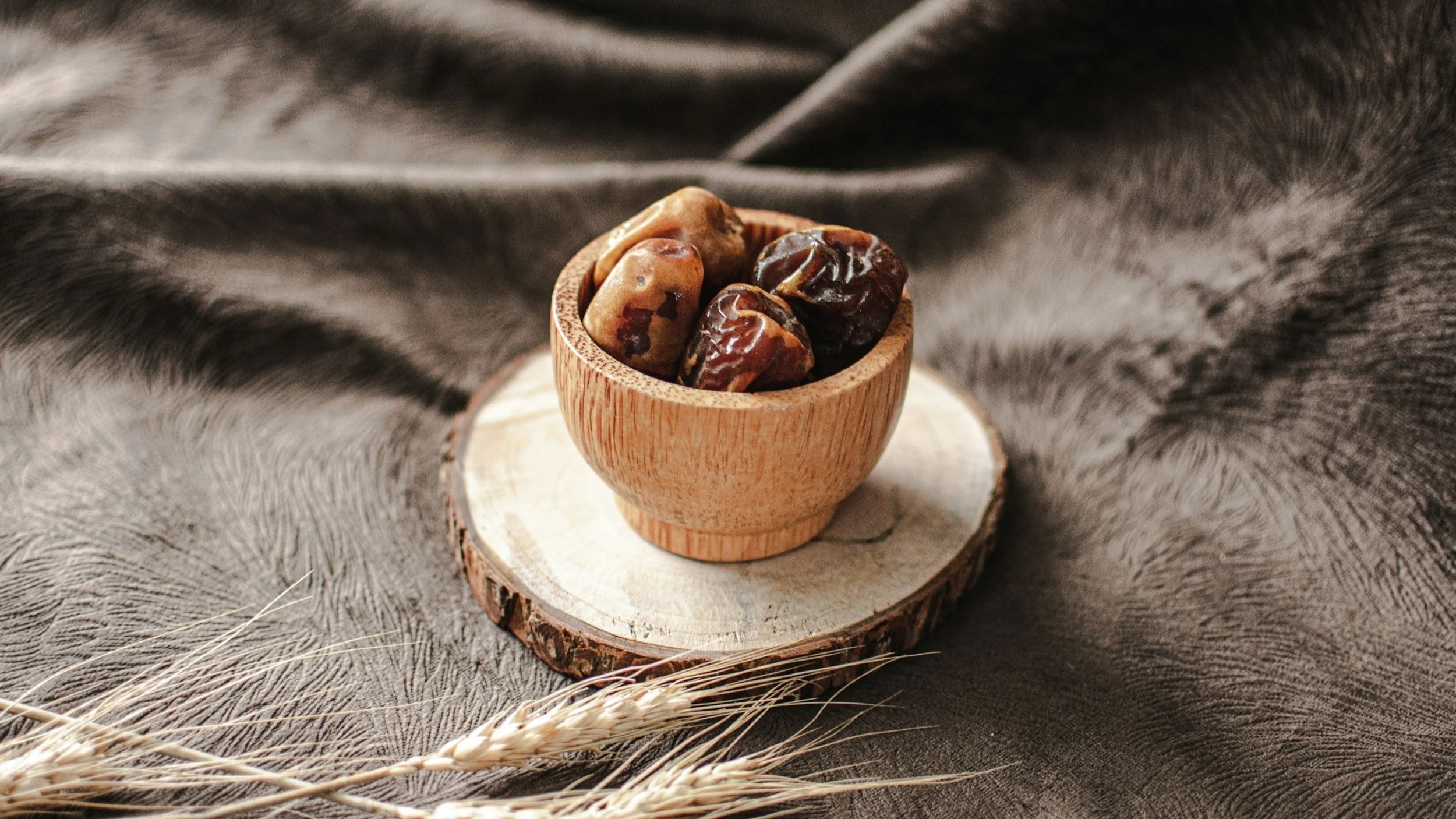 A small wooden cup filled with dates, placed on a cross-section of a tree trunk, with a dark, textured cloth background and some wheat stalks in the foreground.