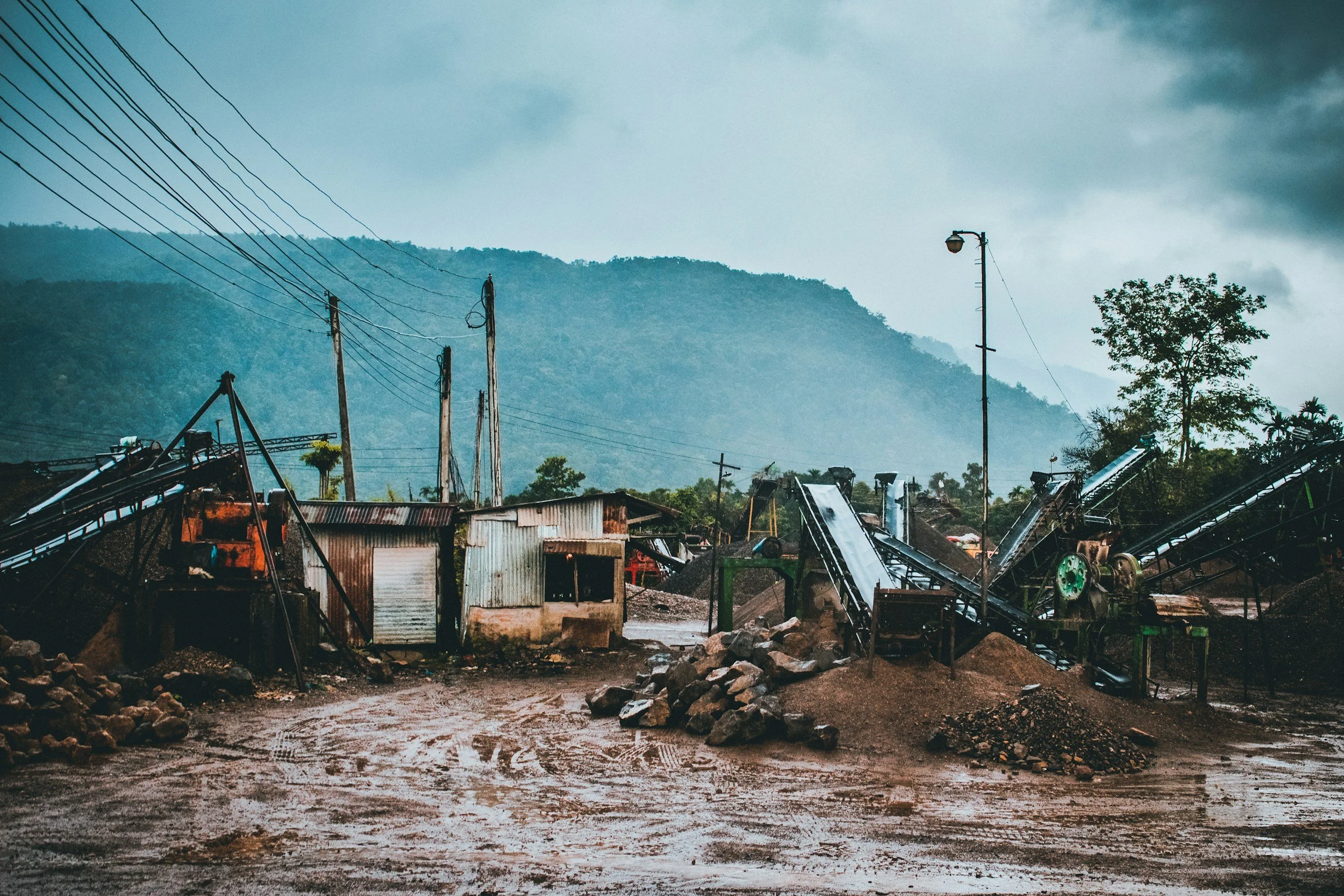 A landscape scene showing a muddy area with debris, damaged structures, and fallen trees. In the background, there are mountains and a cloudy sky, with a row of utility poles and power lines.