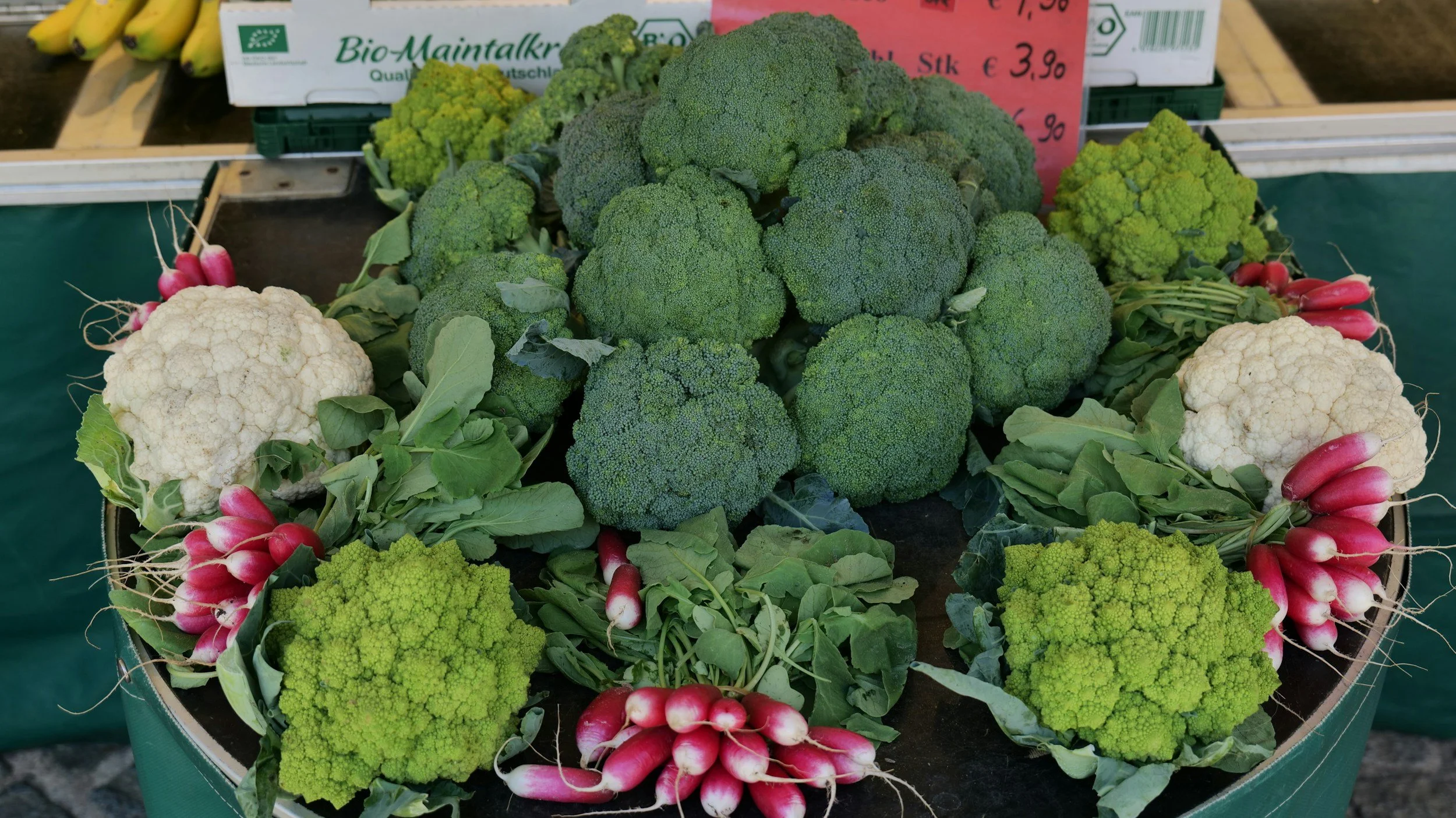 A display of fresh green broccoli, white cauliflower, and red radishes arranged together at a market stall.