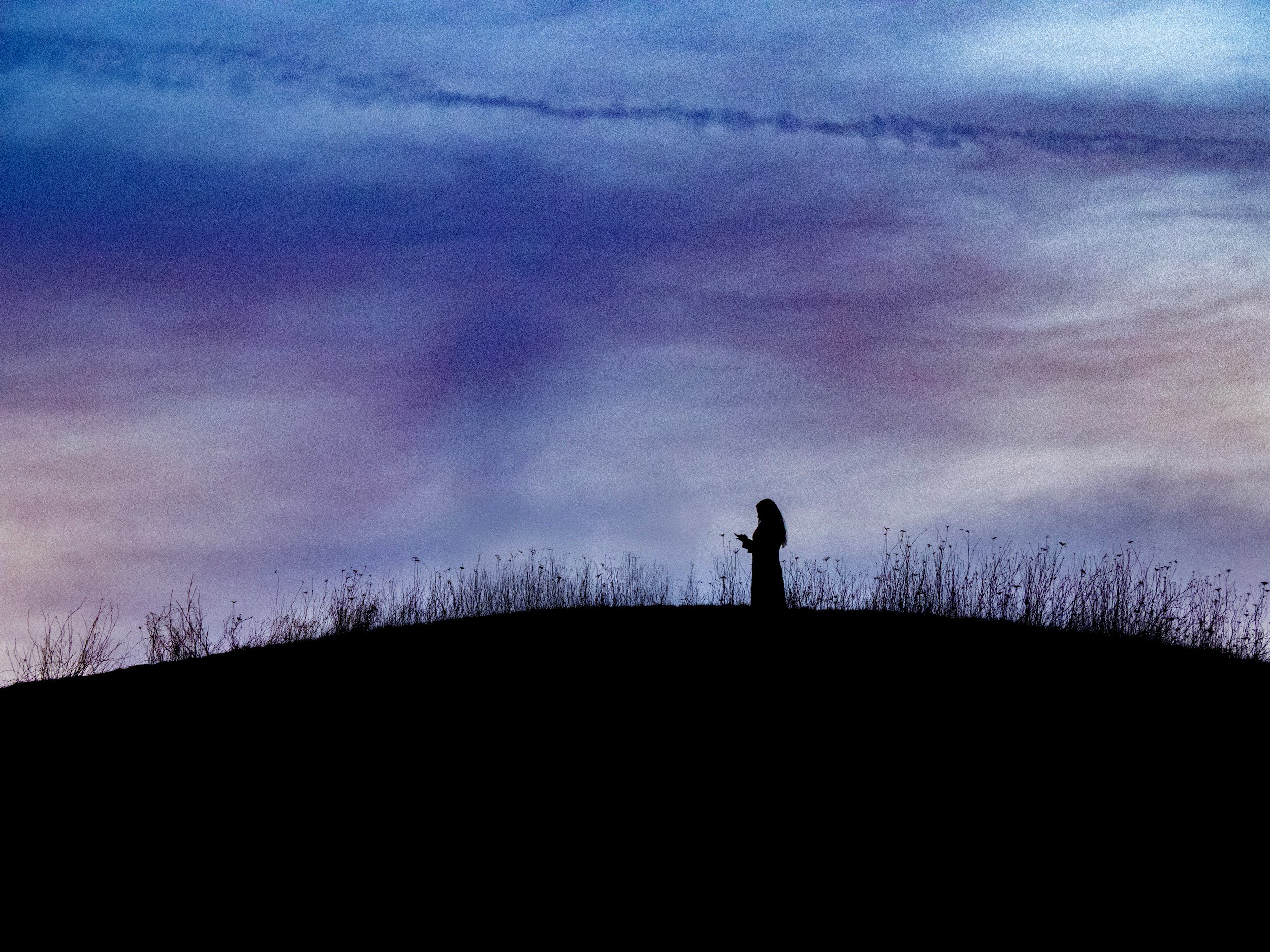 Silhouette of a person standing on a small hill or mound with dry grass, looking at a cloudy twilight sky, with the horizon visible in the distance.