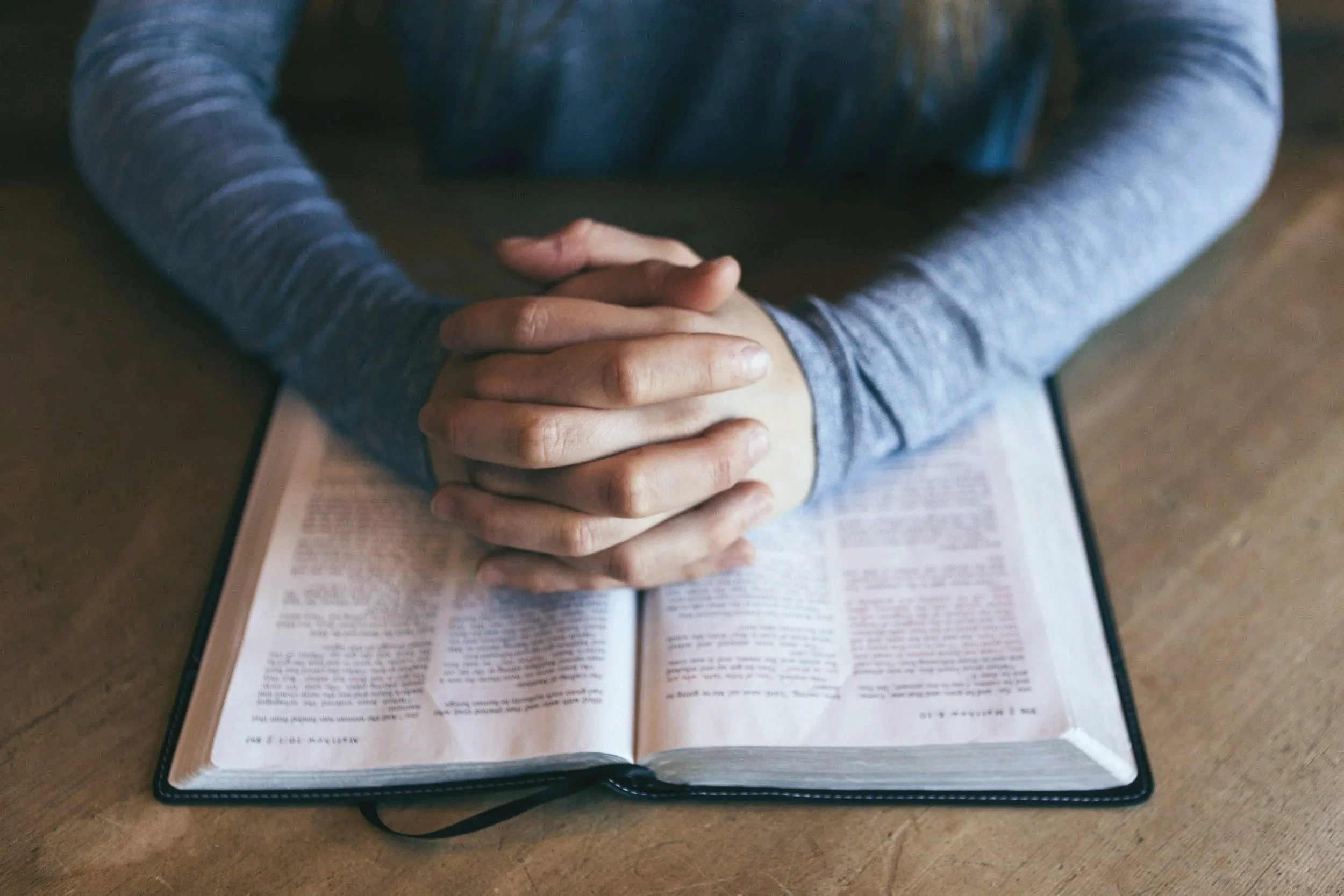 Person sitting at a table with hands clasped over an open book, wearing a long-sleeve gray shirt, the focus is on the hands and book.