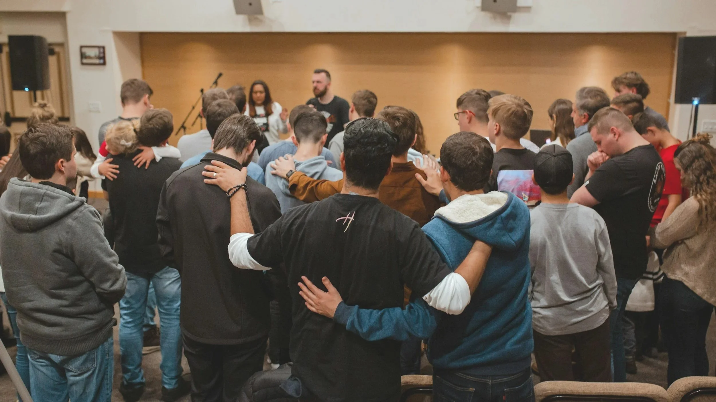 Group of young people standing close together in circle, praying with heads bowed and hands on each other's shoulders in a warmly lit indoor setting.
