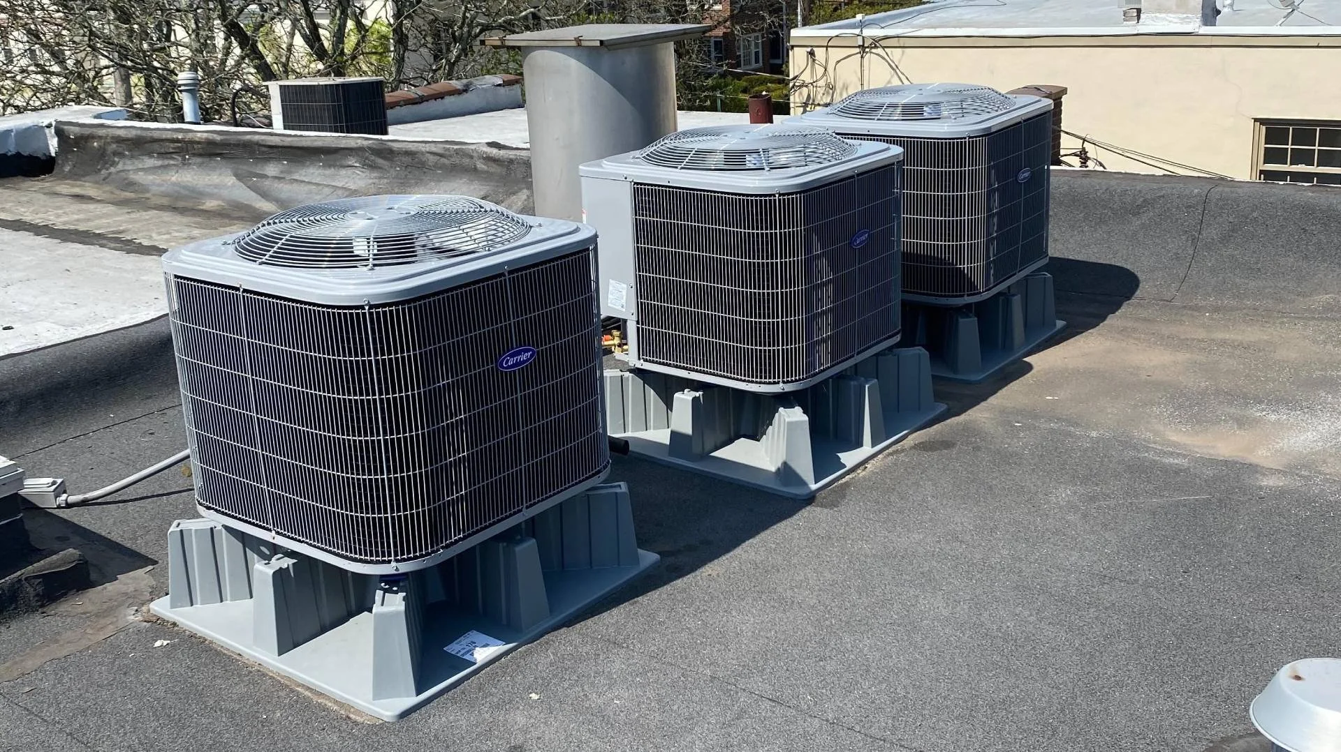 Three Carrier air conditioning units on a rooftop, with a beige building and tree in the background.