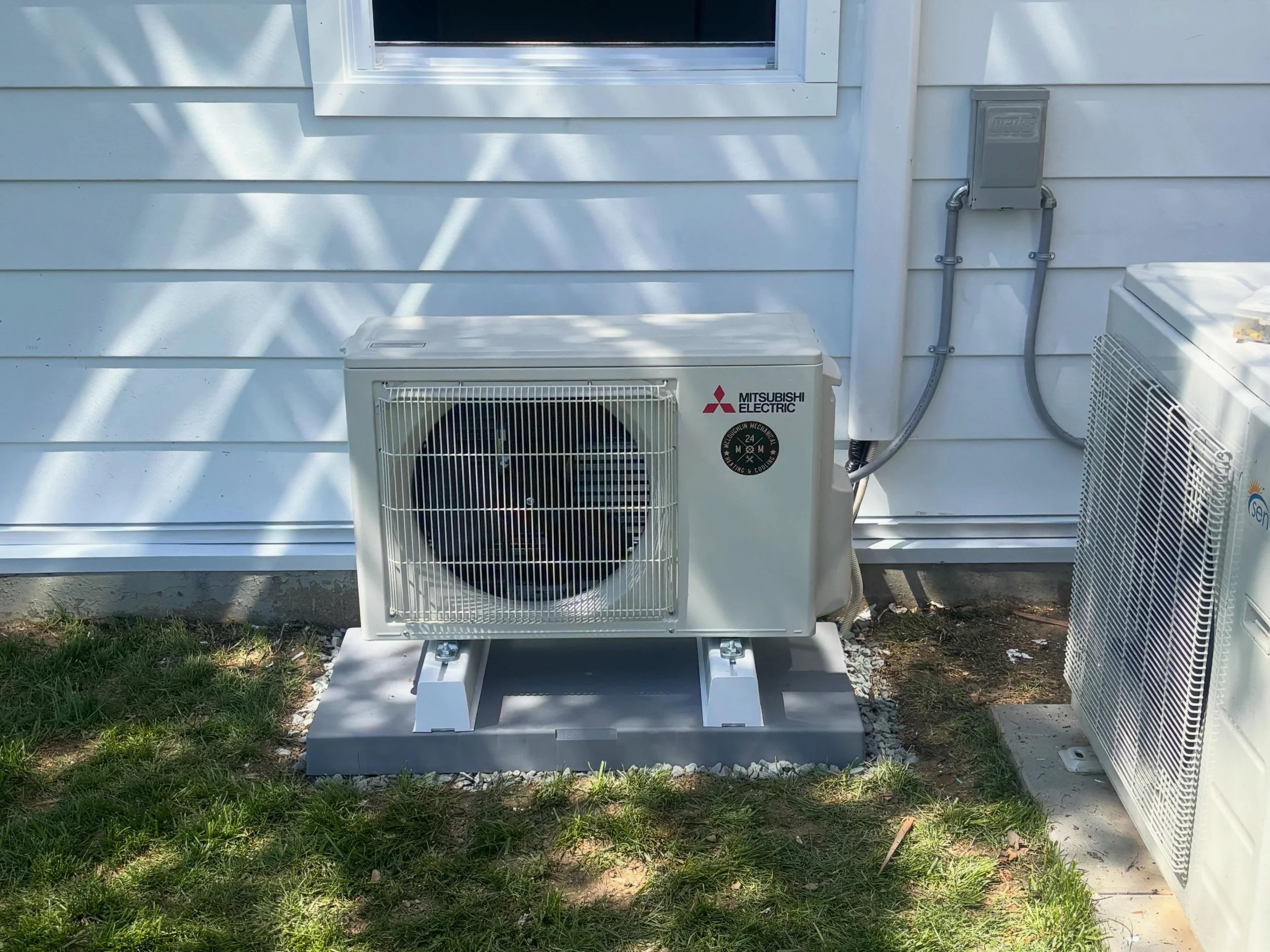 An outdoor white wall with a window, an air conditioning unit labeled 'Mitsubishi Electric', and a gray electrical box with conduit pipes, with part of a lawn and gravel at the bottom.