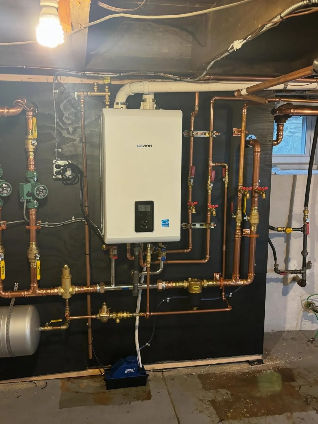 A Navien tankless water heater mounted on a dark wall, with copper pipes and shut-off valves connected to it in a basement setting, including a small window and exposed wiring.