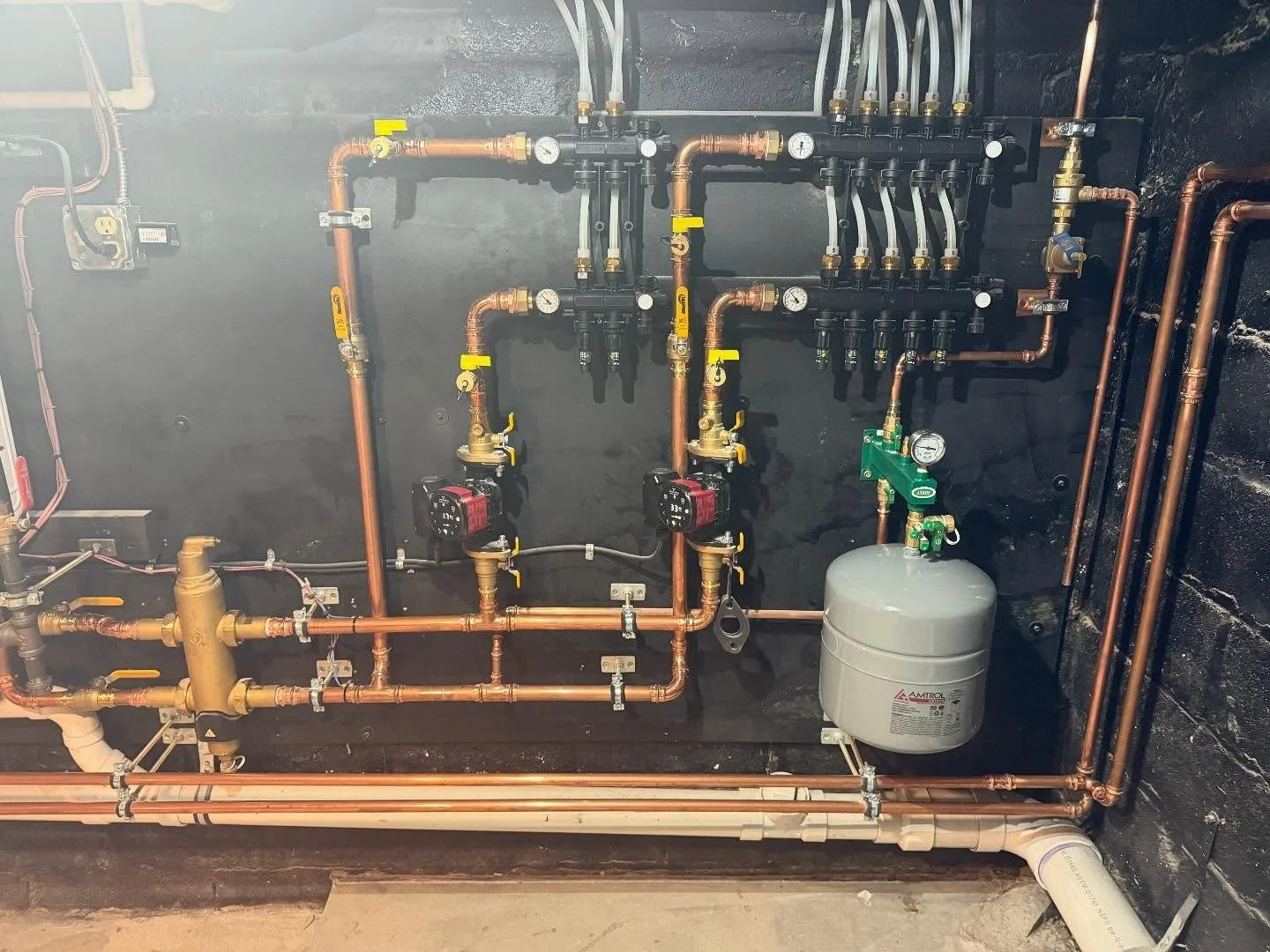 A complex plumbing heating system with copper pipes, gauges, valves, and a gray expansion tank installed on a wall.