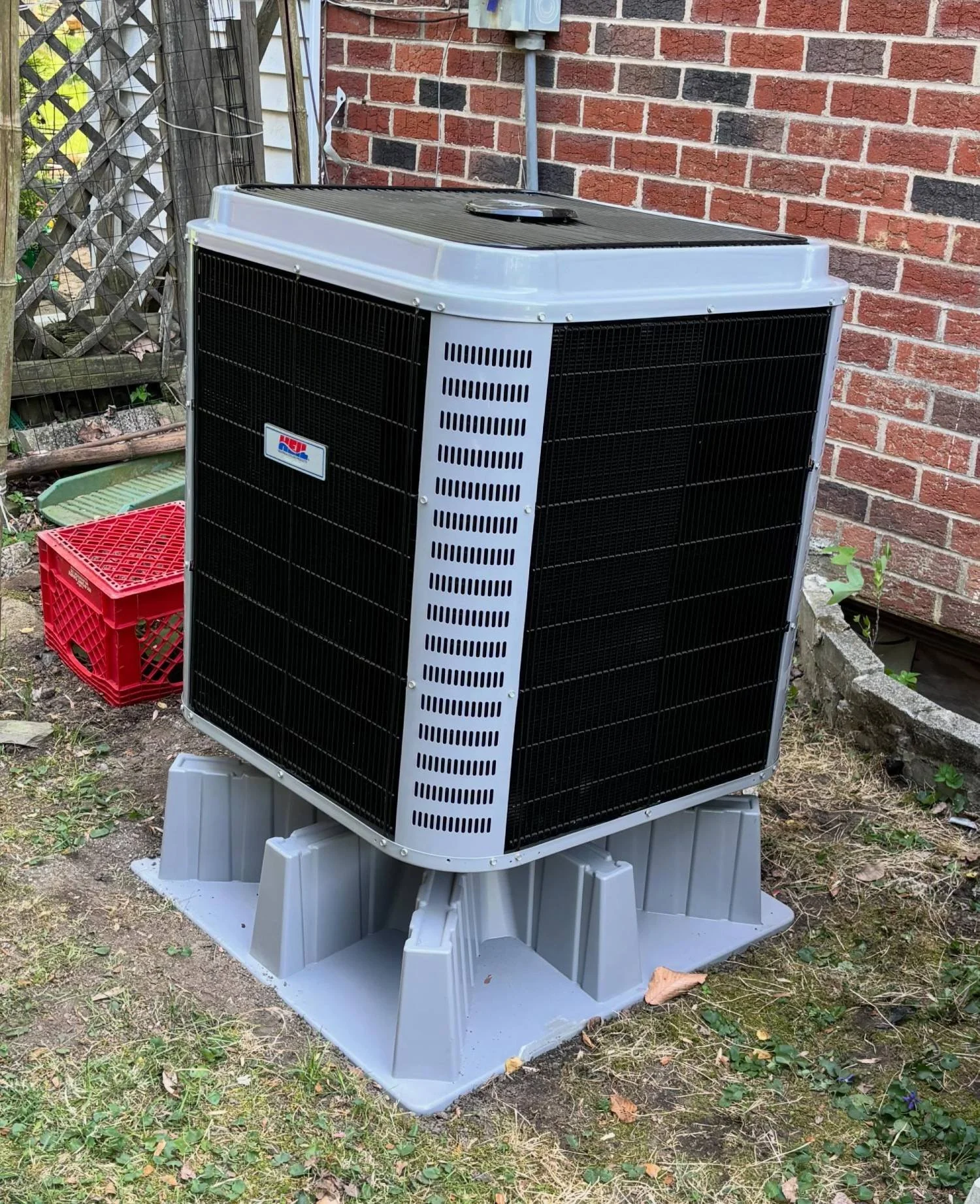 An outdoor air conditioning unit installed on a gray plastic platform next to a brick wall, with a lattice fence and some plants nearby.