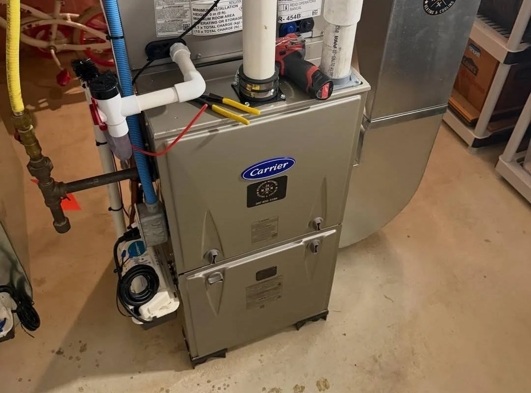A Goodman Carrier furnace with various pipes and a cordless drill on top, located in a utility or laundry room with shelves and storage.