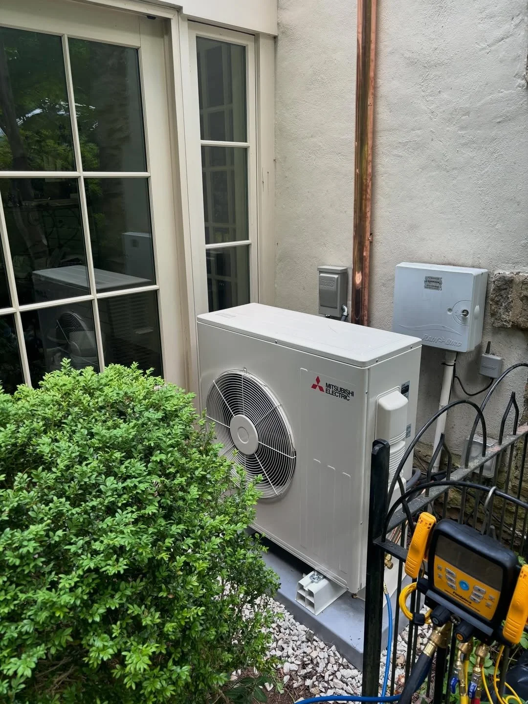 Outdoor view of a Mitsubishi electric air conditioning or heat pump unit installed next to a house wall, with a bush in the foreground and electrical boxes mounted on the wall.