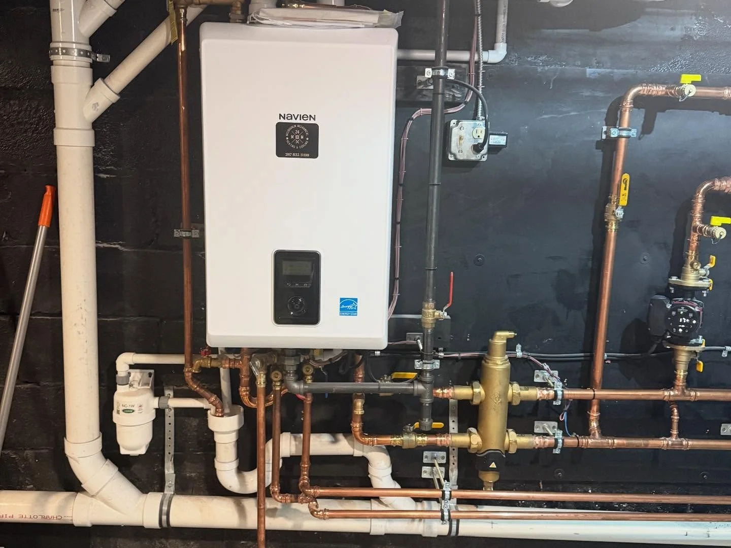 A boiler system with various pipes, valves, and a control panel mounted on a black wall.