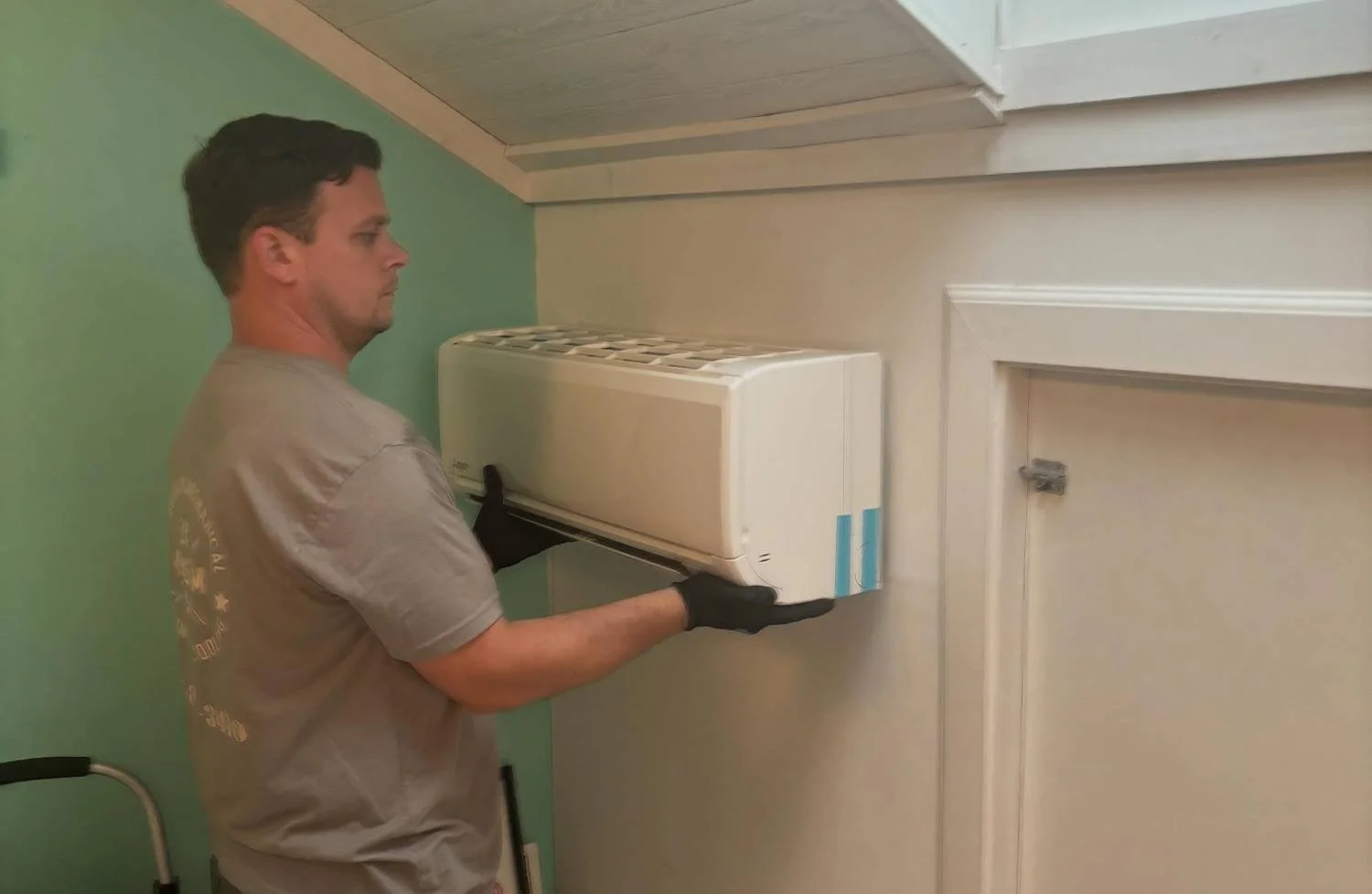 A man wearing a gray t-shirt and black gloves installing or removing an air conditioning unit from a wall.