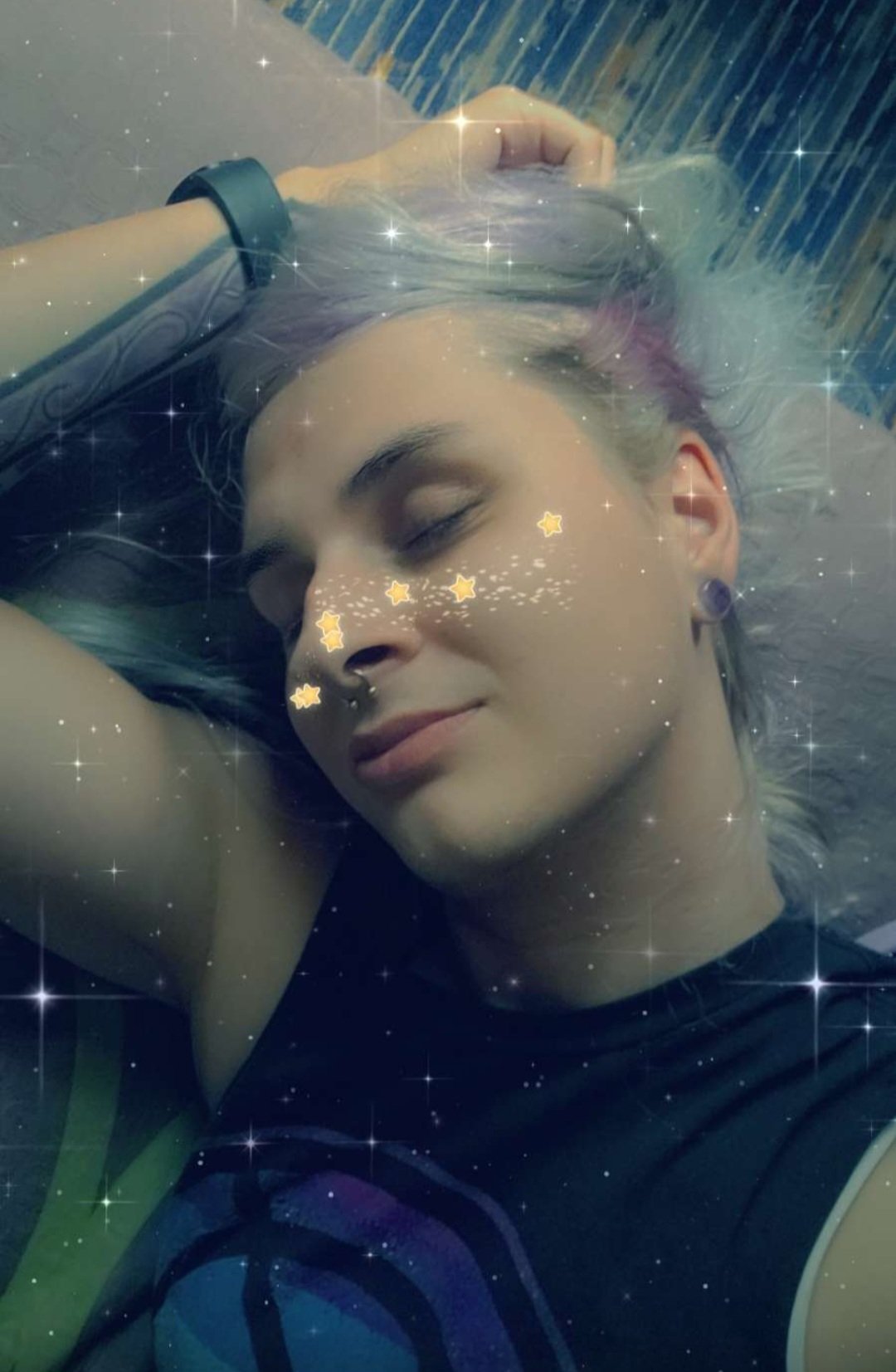 Young woman with blue and pink hair, lying underwater with her eyes closed, smiling gently. There are star-shaped and sparkly effects superimposed on the image.