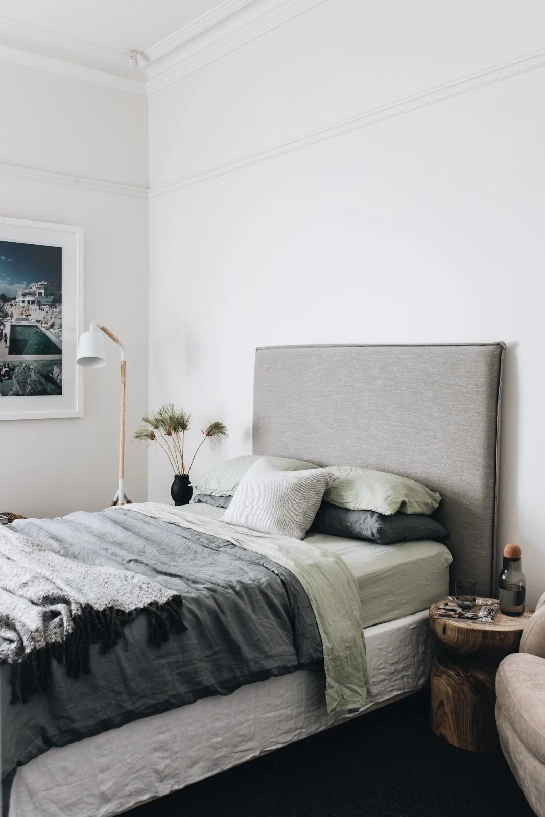 A minimalist bedroom with a beige upholstered headboard, layered gray and cream bedding, a small side table made of carved wood with decor items, a black vase with dried plants, a floor lamp with a white shade, and a framed seaside photograph on the 