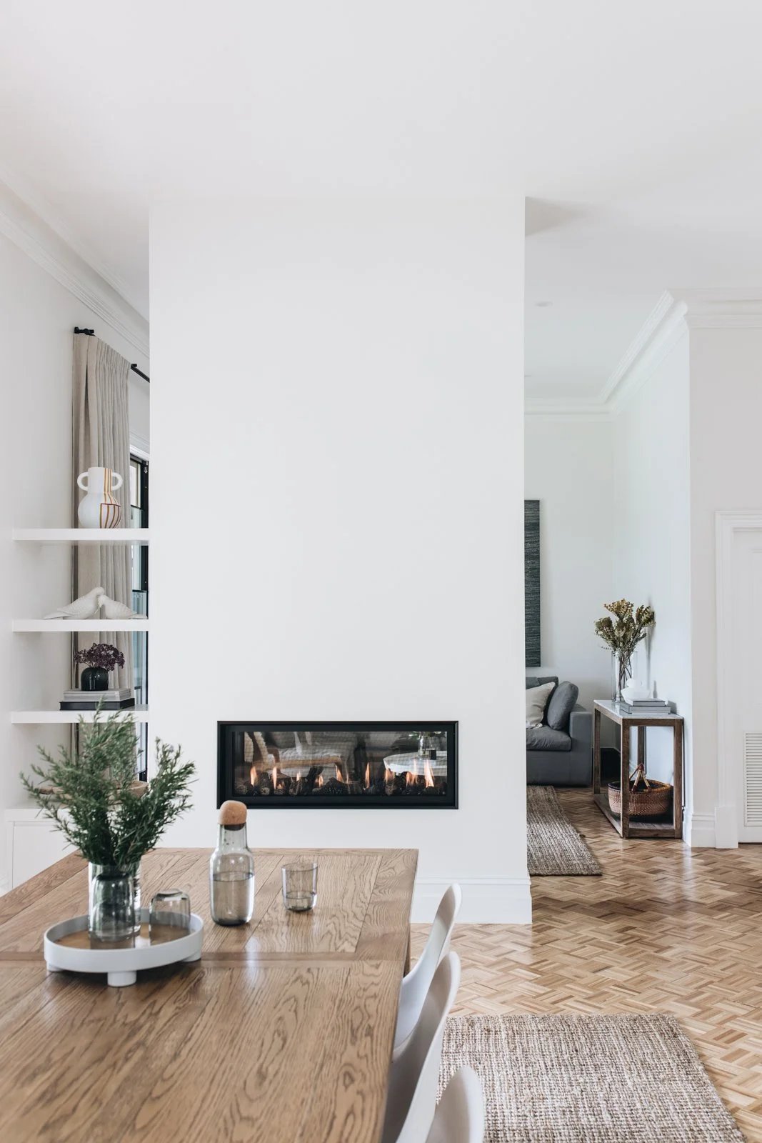 Interior of a modern home with a wooden dining table, a white wall with a built-in fireplace, and a sitting area with a gray sofa and a side table with decor and flowers.
