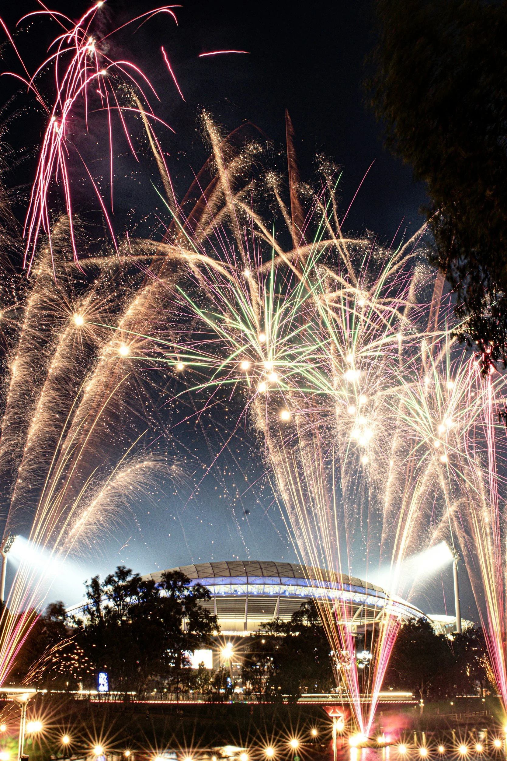 Fireworks exploding above a stadium at night, with trees and lights in the foreground.