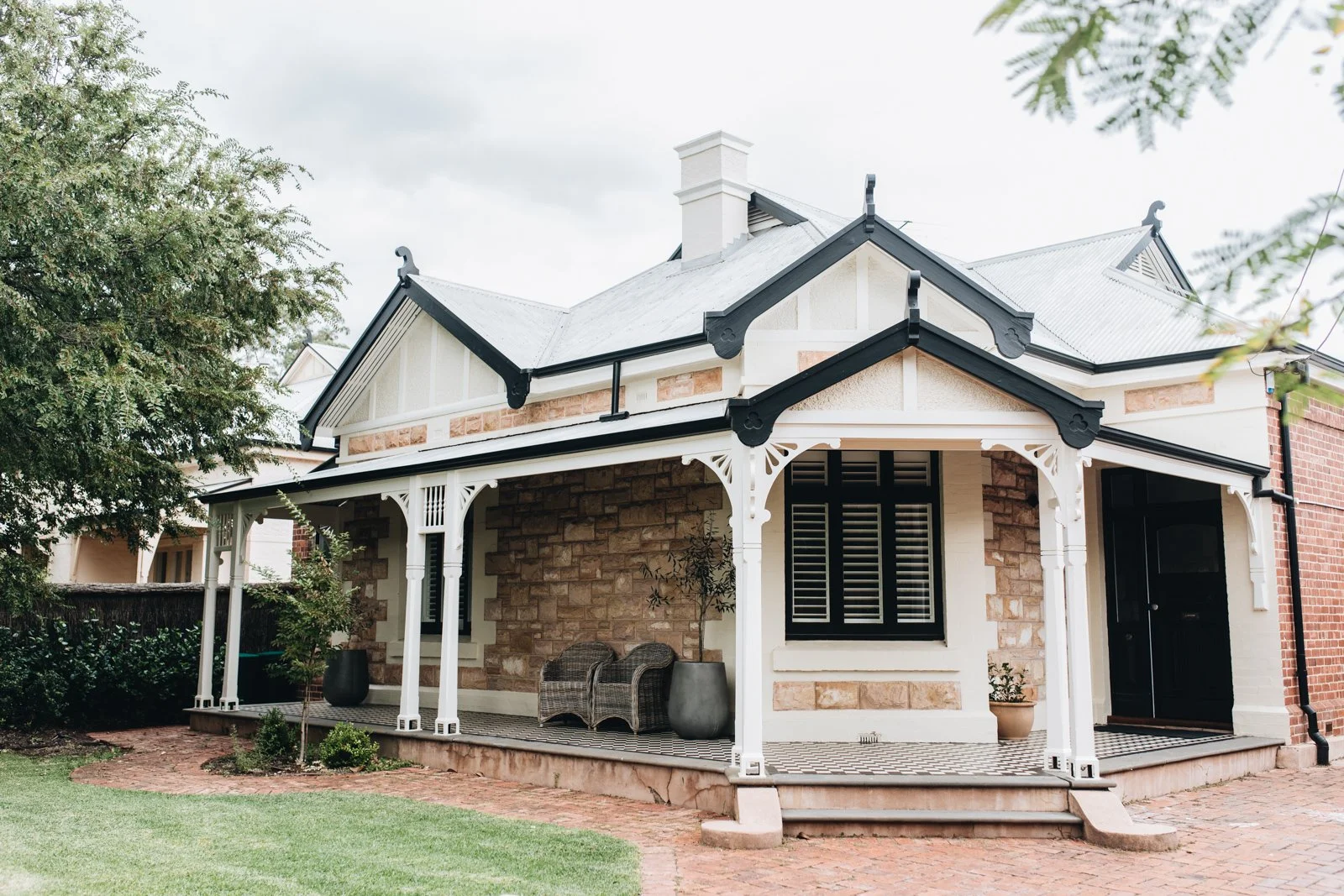 A charming house with a decorative porch, white trim, brick and stone exterior, and a patterned tile porch area surrounded by greenery.