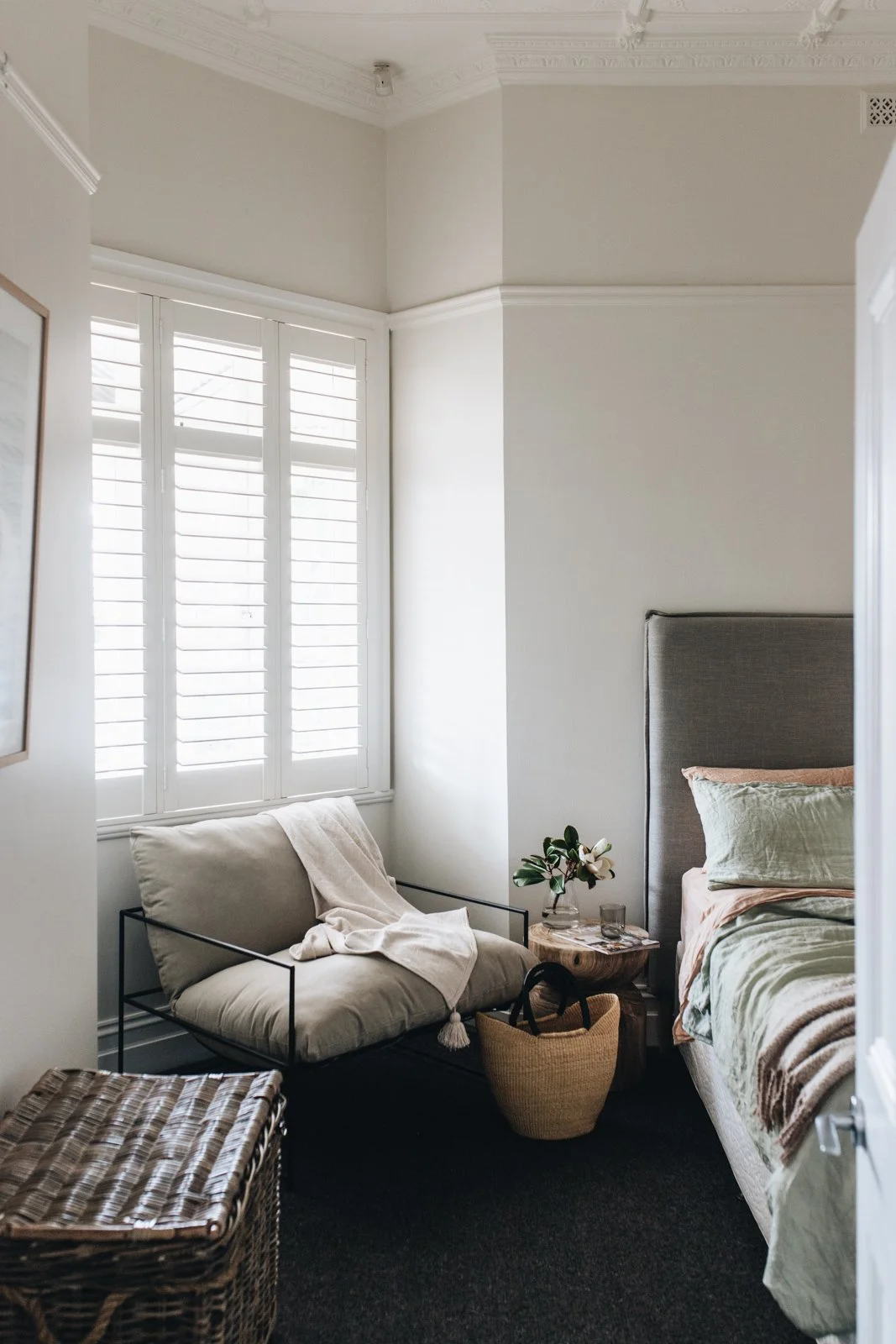 A cozy bedroom corner with a bed, gray upholstered headboard, and green and pink bedding. Next to the bed, a small wooden side table holds a flower vase, a glass, and some books. A beige armchair with a throw blanket, a basket, and a woven ottoman ar