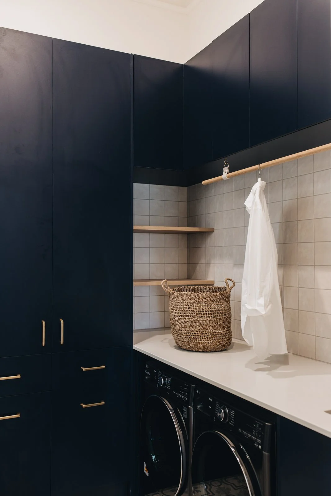 A laundry room with navy blue cabinets and shelves, beige tiled wall, a woven basket on a countertop, and front-loading washing machines.