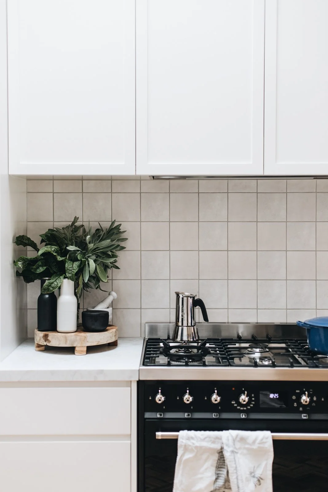 Kitchen with white cabinets, tiled beige backsplash, green plant in black and white vases on a corner shelf, and a stove with a kettle.