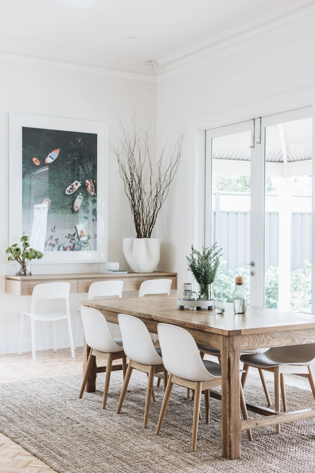 Bright dining room with a wooden table, white chairs, and a large window. Decor includes plants, a large white vase with tall branches, and wall art with boats.