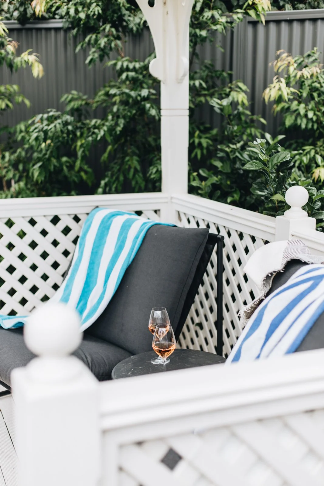 A cozy outdoor patio with dark gray cushioned chairs, blue and white striped towels or blankets, and two glasses of rosé wine on a small round table. There is a white lattice fence and lush green bushes in the background.