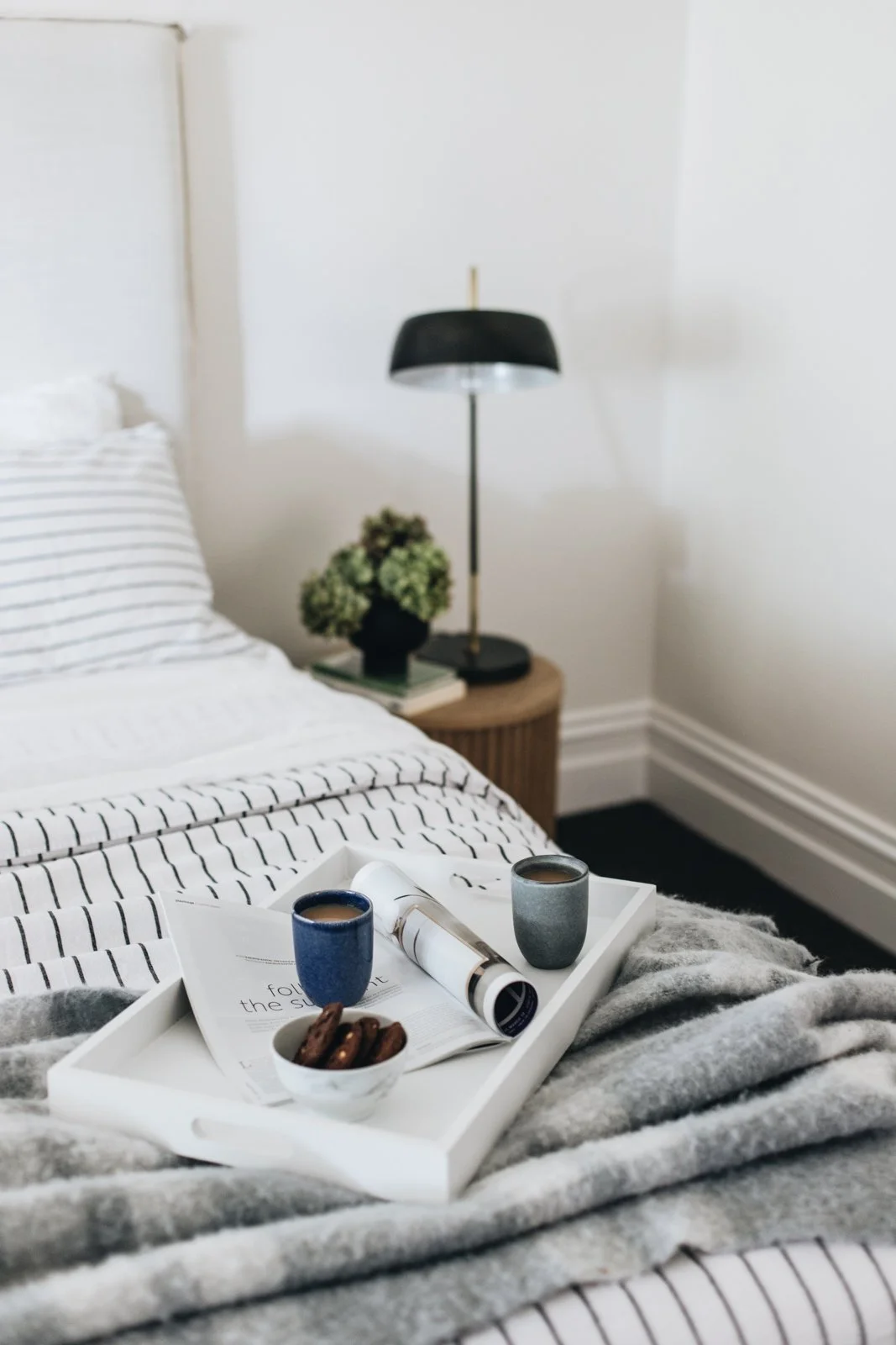 A bedroom scene with a bed, a nightstand with a black lamp, a small potted plant, a magazine, and a tray with two cups of coffee and a bowl of cookies.