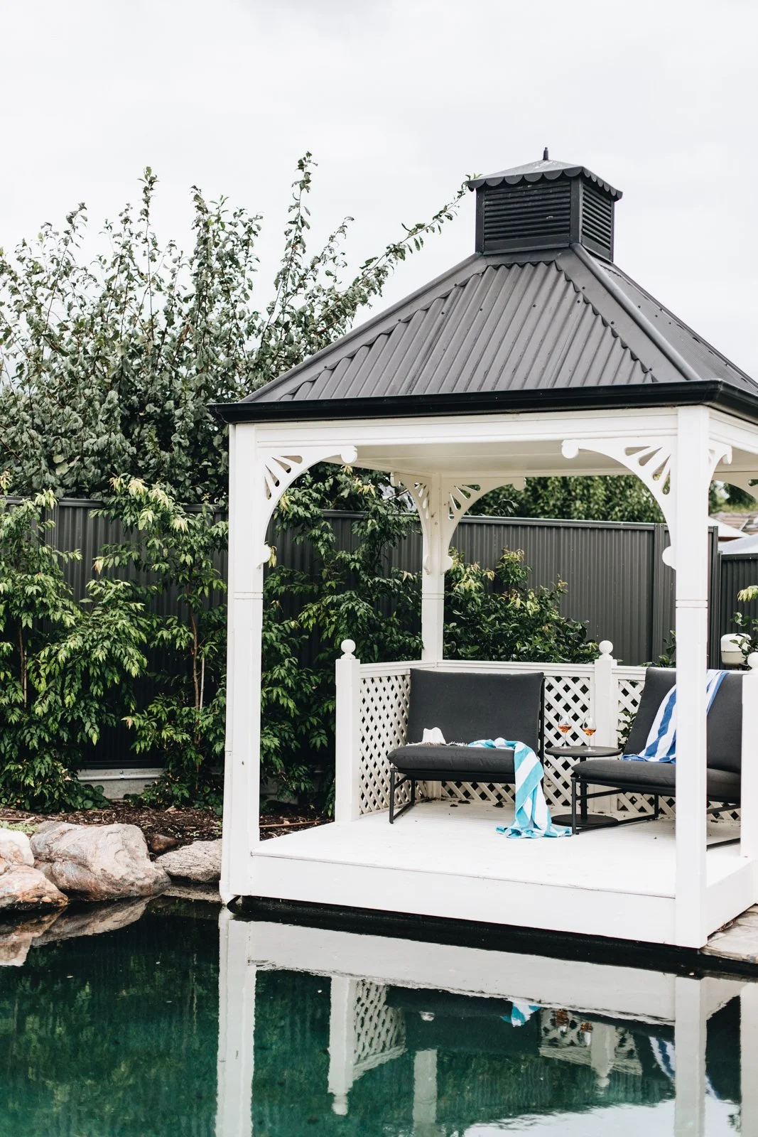A white gazebo by a pond with two black outdoor chairs and some towels and drinks, surrounded by greenery.