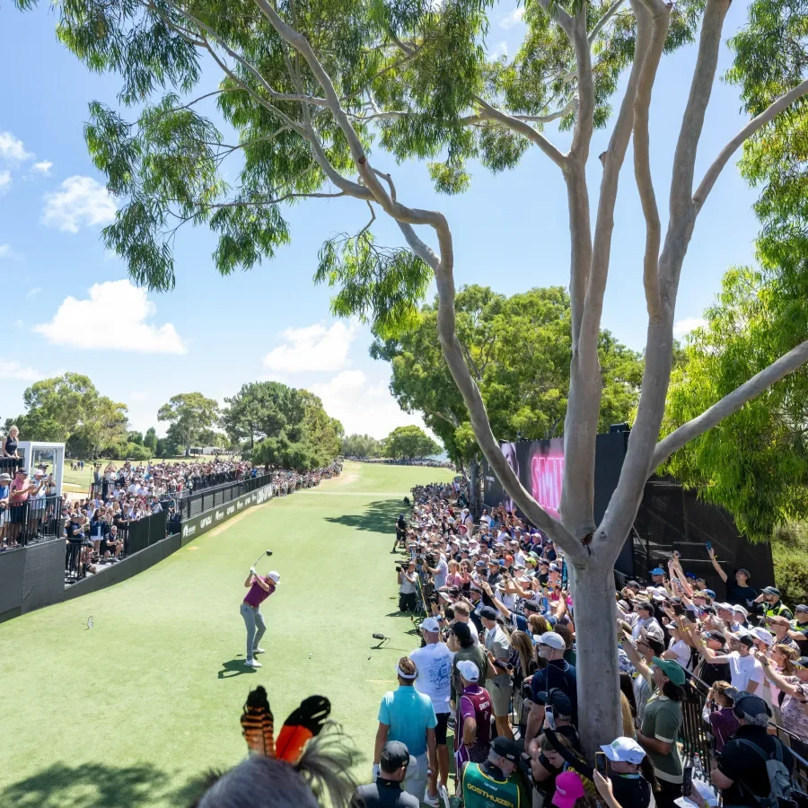 A golfer in a maroon shirt and gray pants swinging a club on a lush green golf course surrounded by a large crowd of spectators under a clear blue sky.