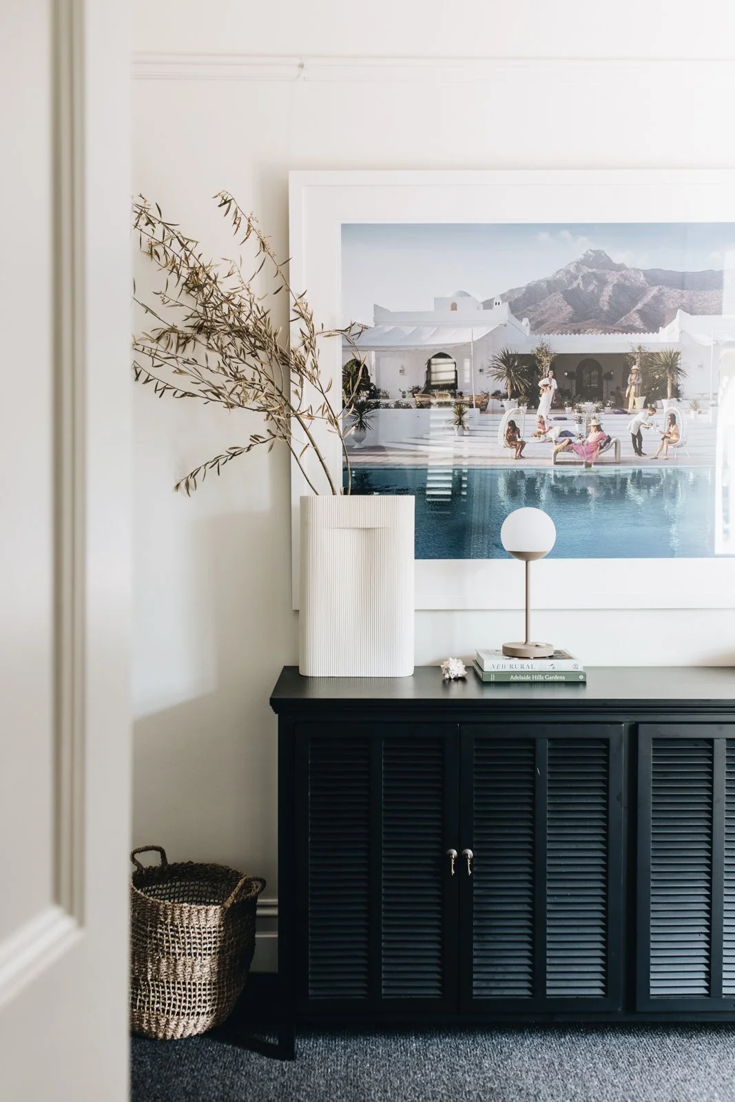 Interior decor with a black cabinet, white decor items, a small table lamp, and a large framed artwork of a poolside scene with mountains in the background.
