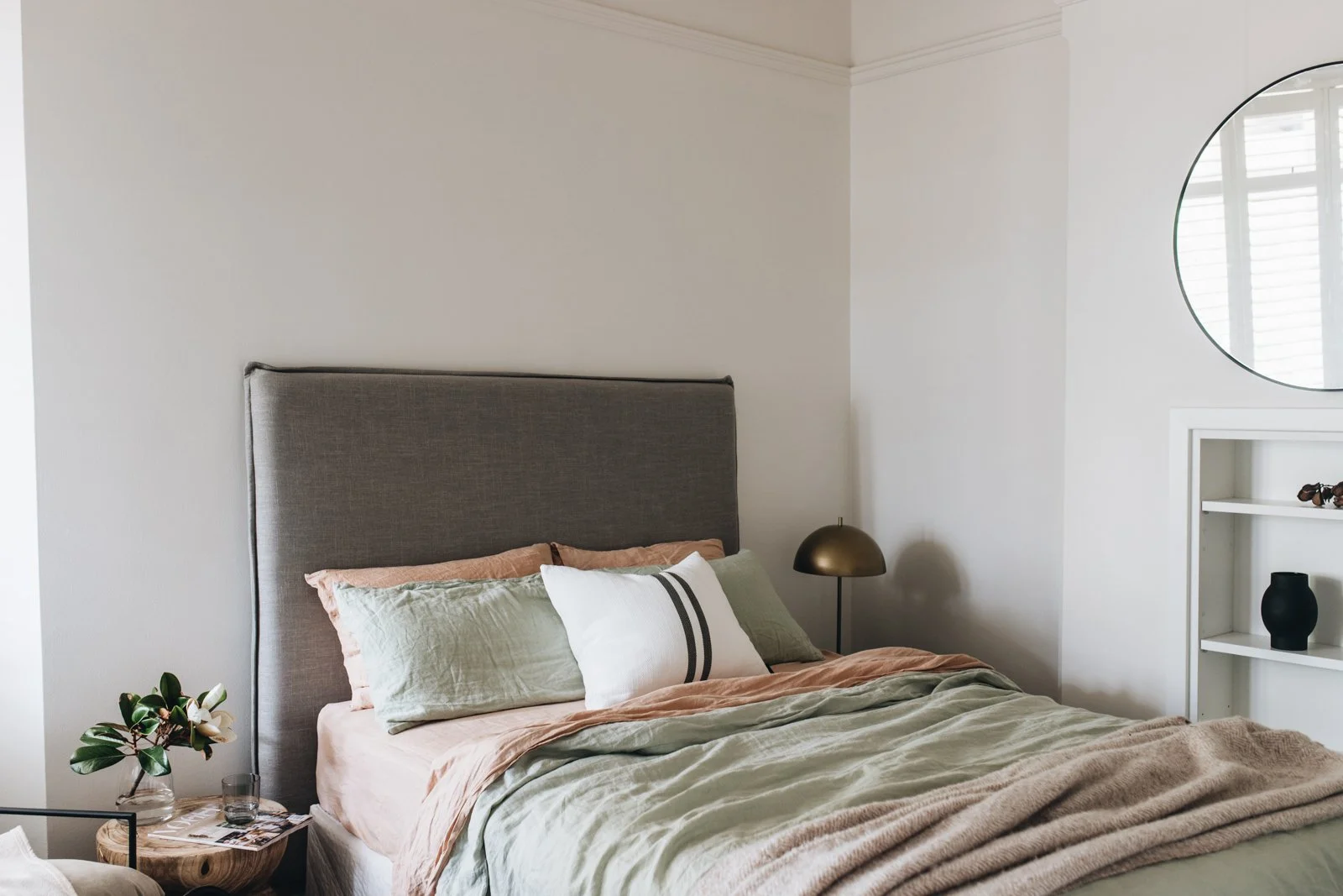 A modern bedroom featuring a gray upholstered headboard, neutral-toned bedding with multiple pillows, a black bedside lamp, a small side table with a plant and glass, and a white built-in shelf with decorative items. A circular mirror is on the wall 