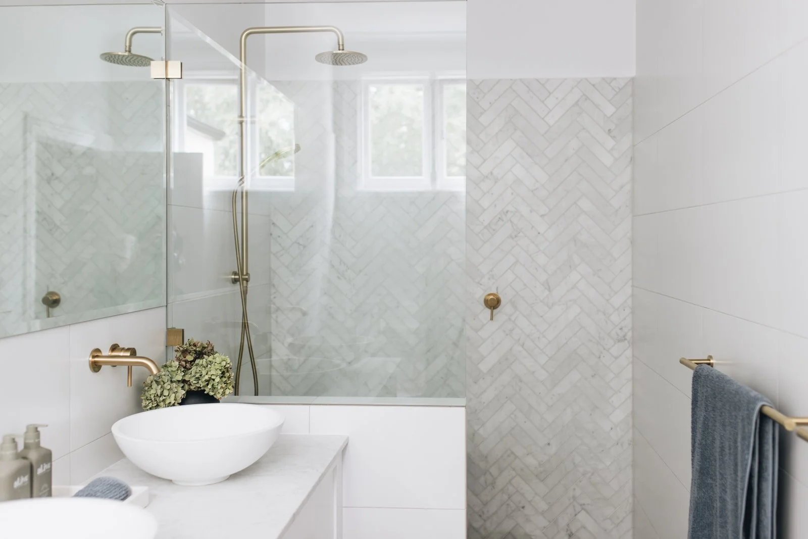 Modern bathroom with white marble and tile walls, glass shower enclosure, white vessel sink, gold fixtures, and a towel bar with a blue towel.