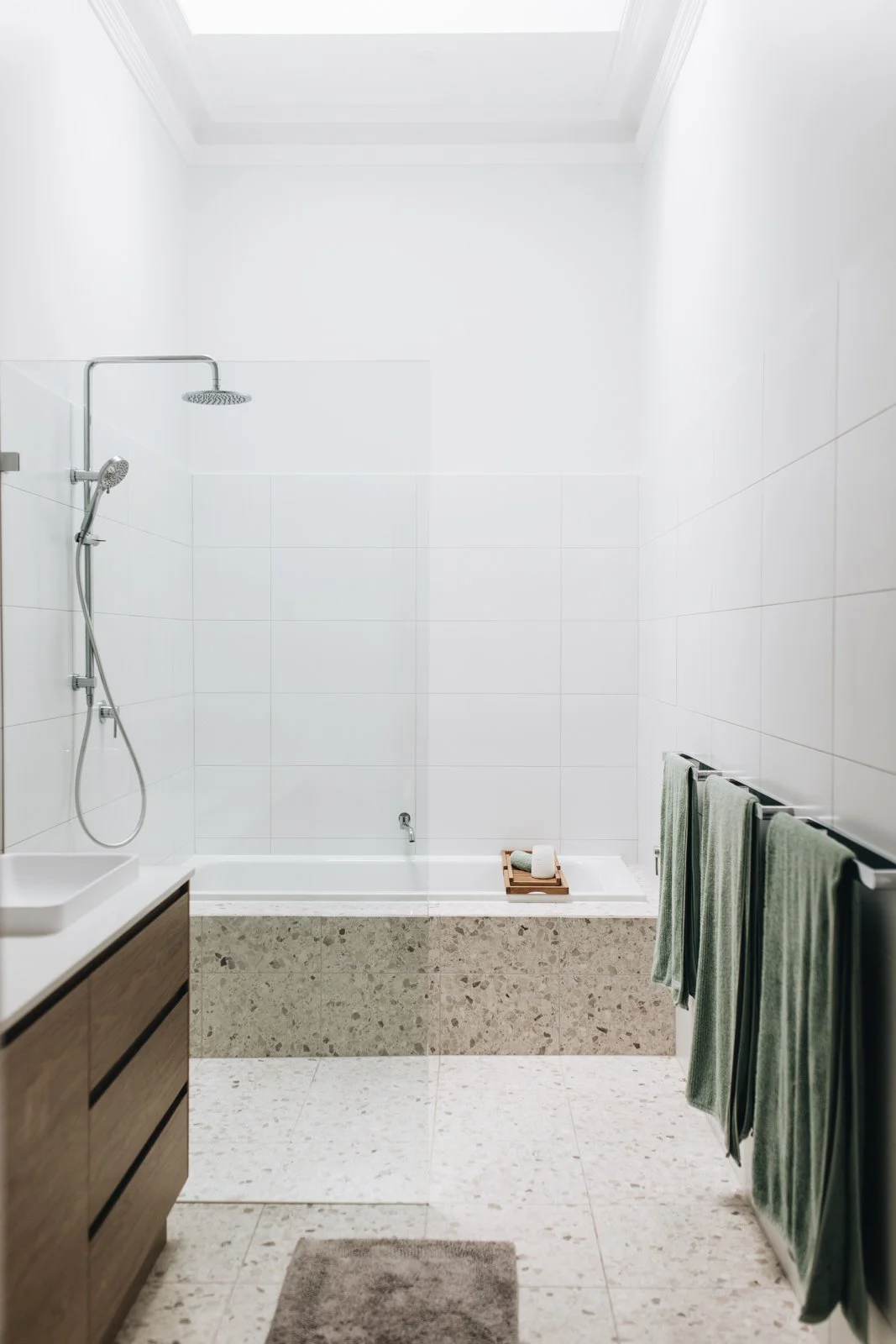 Minimalist bathroom with white tiled walls, a bathtub, a wooden vanity with a white basin, a towel rack with green towels, and a small gray rug on a speckled tile floor.