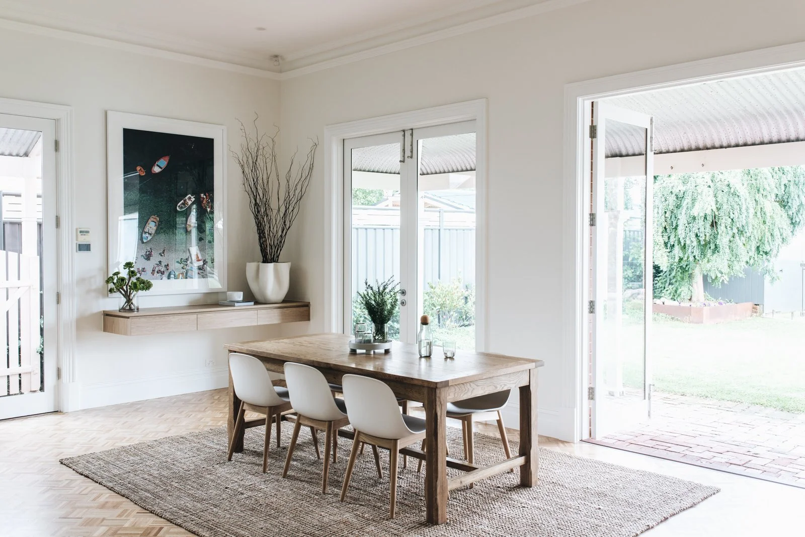 Dining room with wooden table, six white chairs, glass door leading outside, wall with artwork and decorative plant, and a rectangular rug on light hardwood floor.