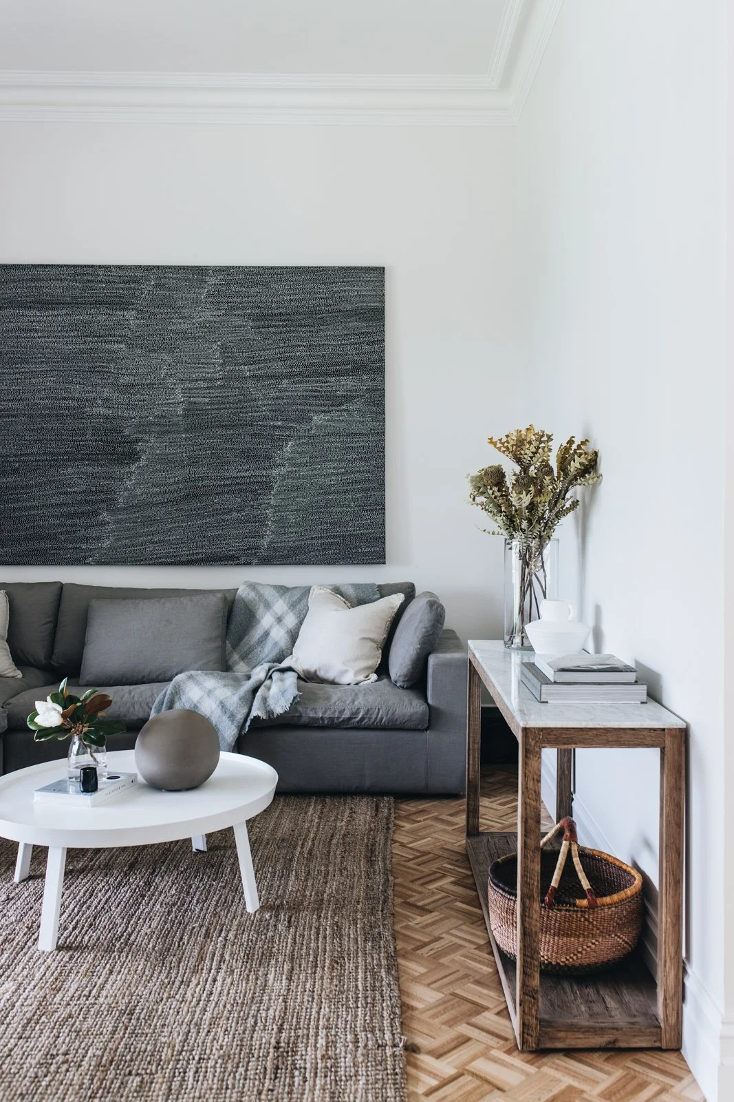 Living room with a gray sectional sofa, a round white coffee table, and a wooden side table with books, vases, and dried flowers. There is a large textured dark art piece on the wall above the sofa, and the floor is a patterned wood with a brown wove