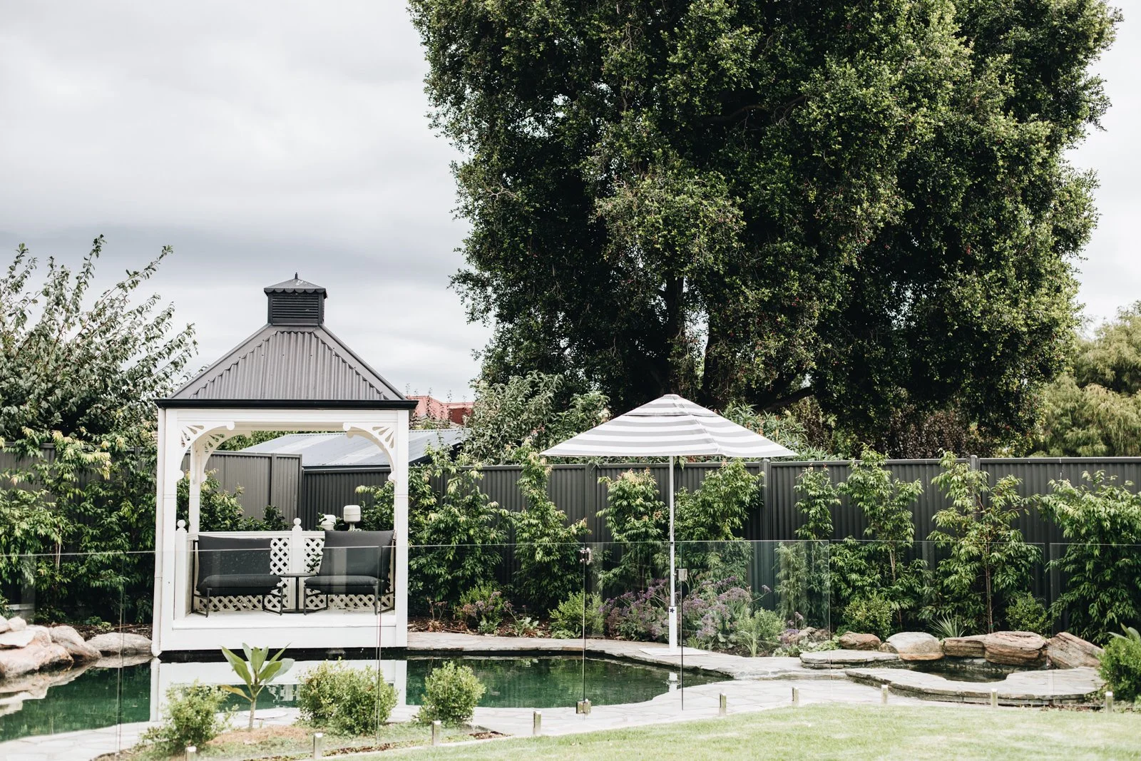 A backyard garden with a pond, a white gazebo, a striped umbrella, and lush green trees and plants.