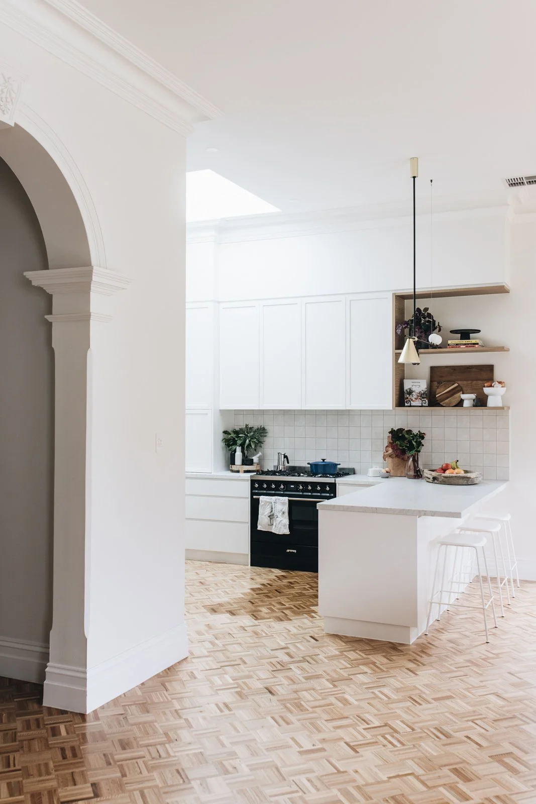 Modern white kitchen with wooden shelving, potted plants, and a black stove, featuring a white countertop and barstools.