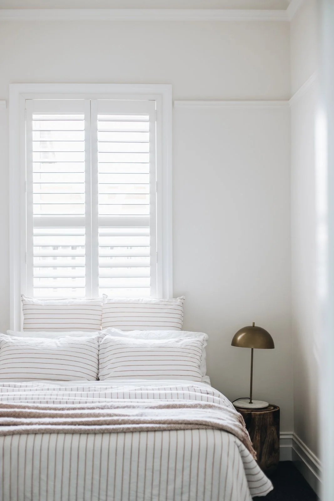 A bedroom with white walls, a window with white shutters, a bed with white and beige striped bedding and pillows, and a small bedside table with a gold-colored lamp.