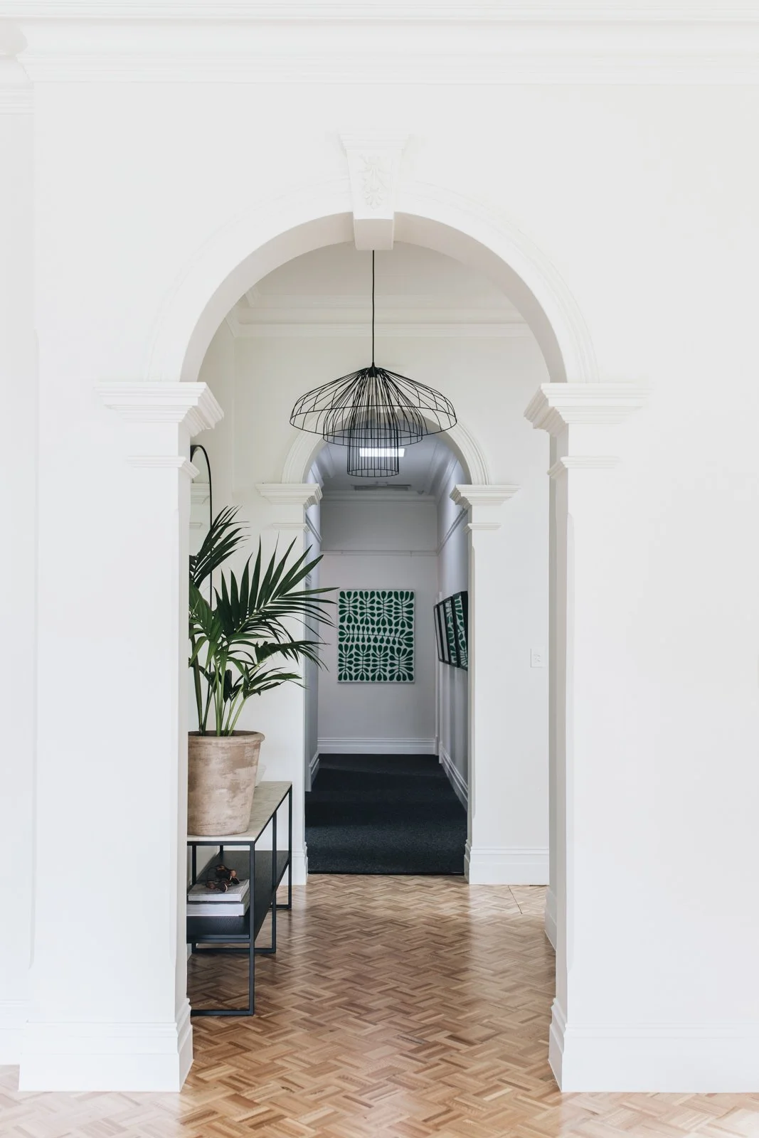 Interior hallway with white arches, black wire pendant light, potted plant, black console table, and framed artwork on the walls.