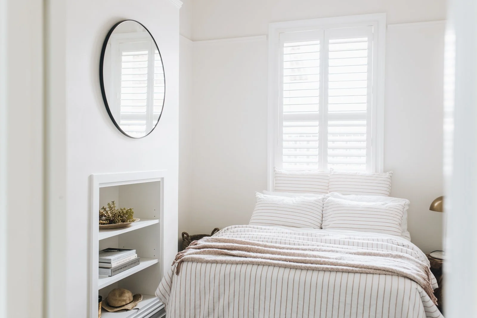 A bright bedroom with white walls, a bed with striped bedding, two pillows, and a blanket. There is a large window with white shutters, a round mirror on the wall, and a white shelf with books, plants, and decorative items.