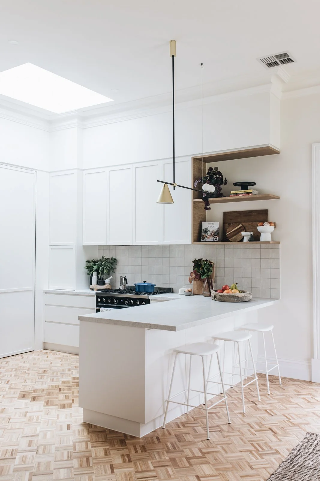 Minimalist white kitchen with wooden accents, open shelving, potted plants, and a light fixture with a black rod and gold cone-shaped shade.